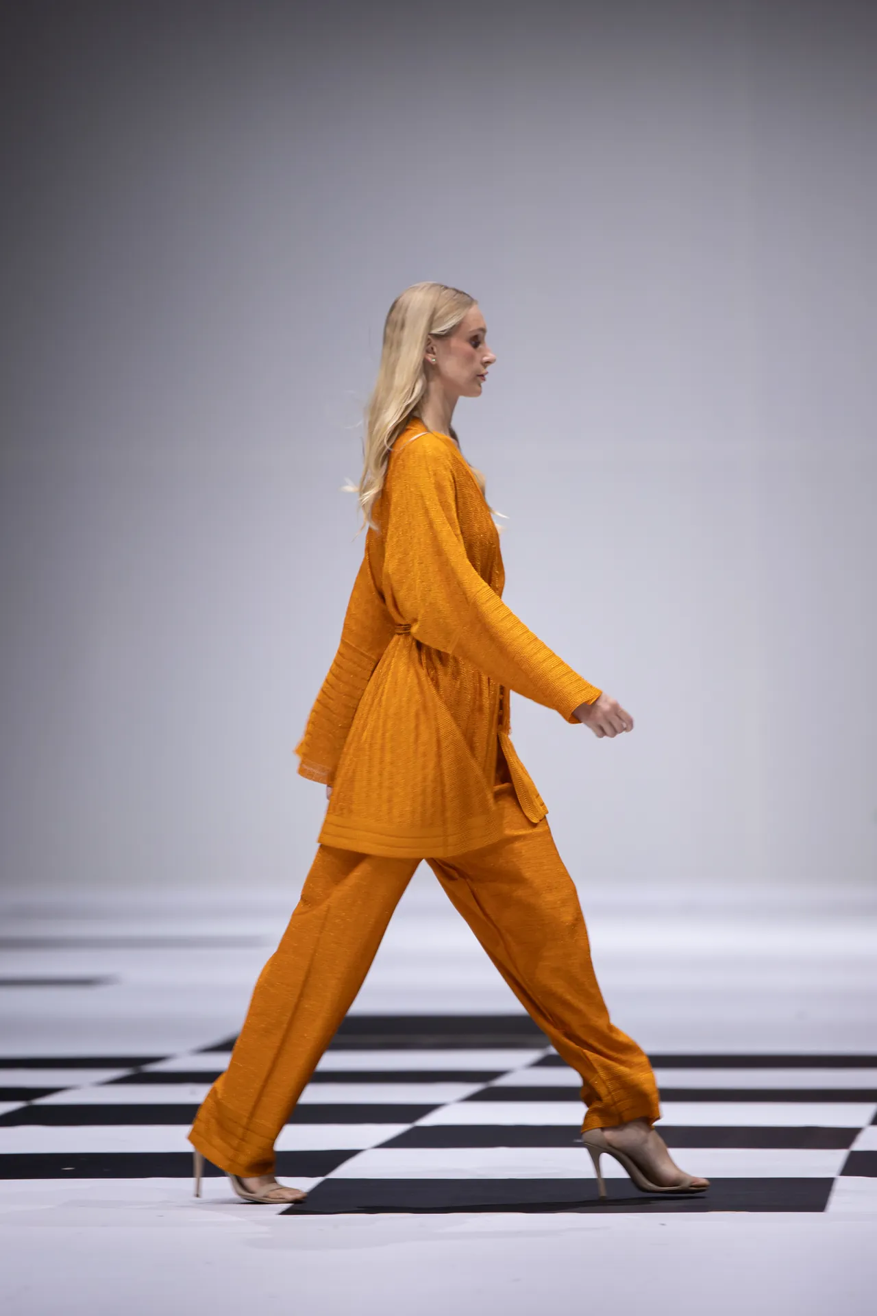 Female model walking on a fashion runway in a bright orange coordinated outfit against a gray backdrop, viewed from the side.