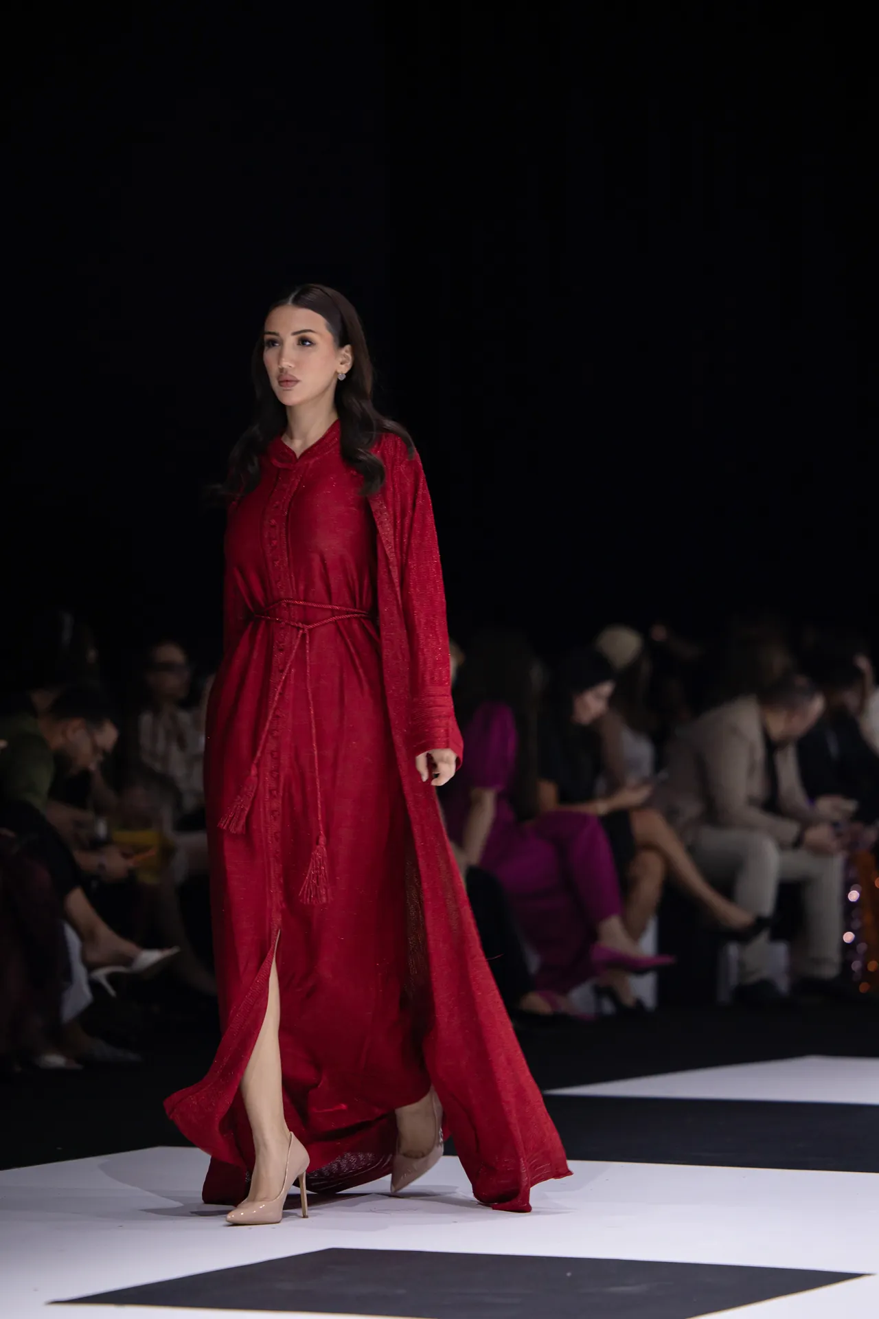 Female model in a red floor-length dress walking a runway, audience blurred in the background.