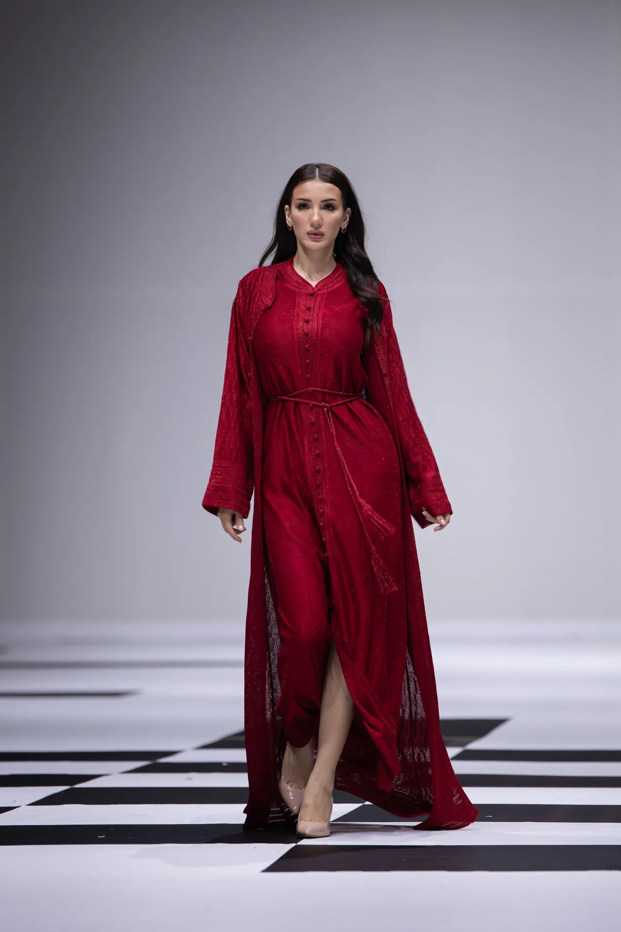 Model walking down a runway in a long red dress with a sash, beige heels, on a black-and-white checkered floor.