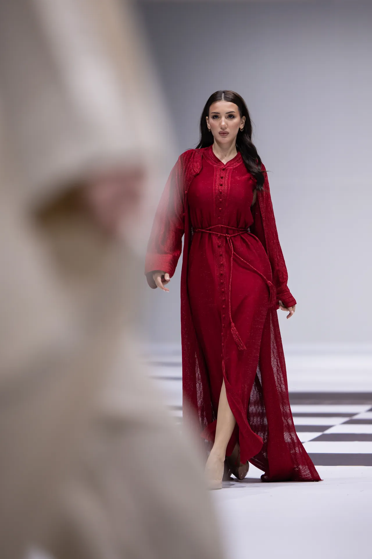 Female runway model in a crimson, floor-length dress walks toward the camera on a black-and-white checkered runway; blurred foreground spectator on the left.