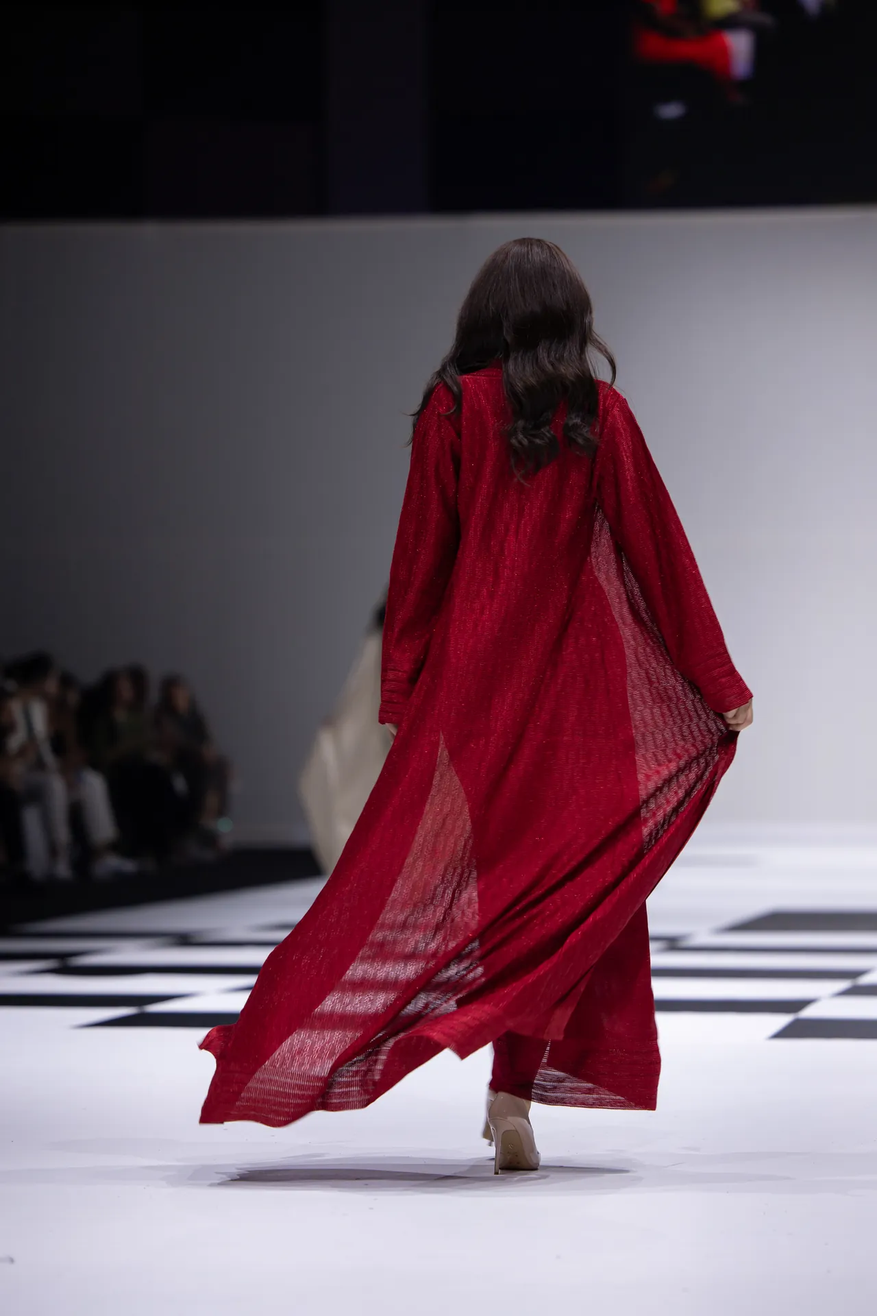 Back-view of a model walking a fashion show in a long red sheer gown, fabric flowing behind on a black-and-white checkered runway.