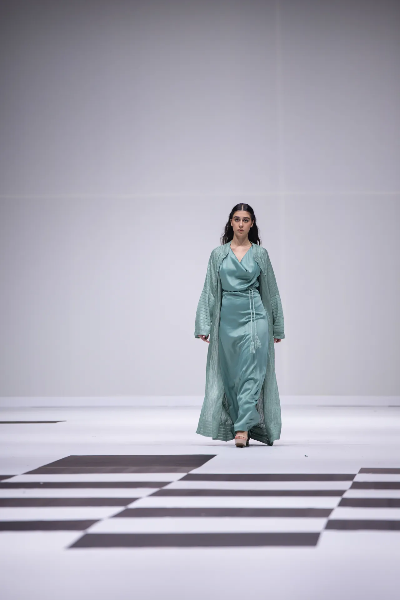Model walking on a minimalist runway in a teal satin dress with a matching sheer robe.