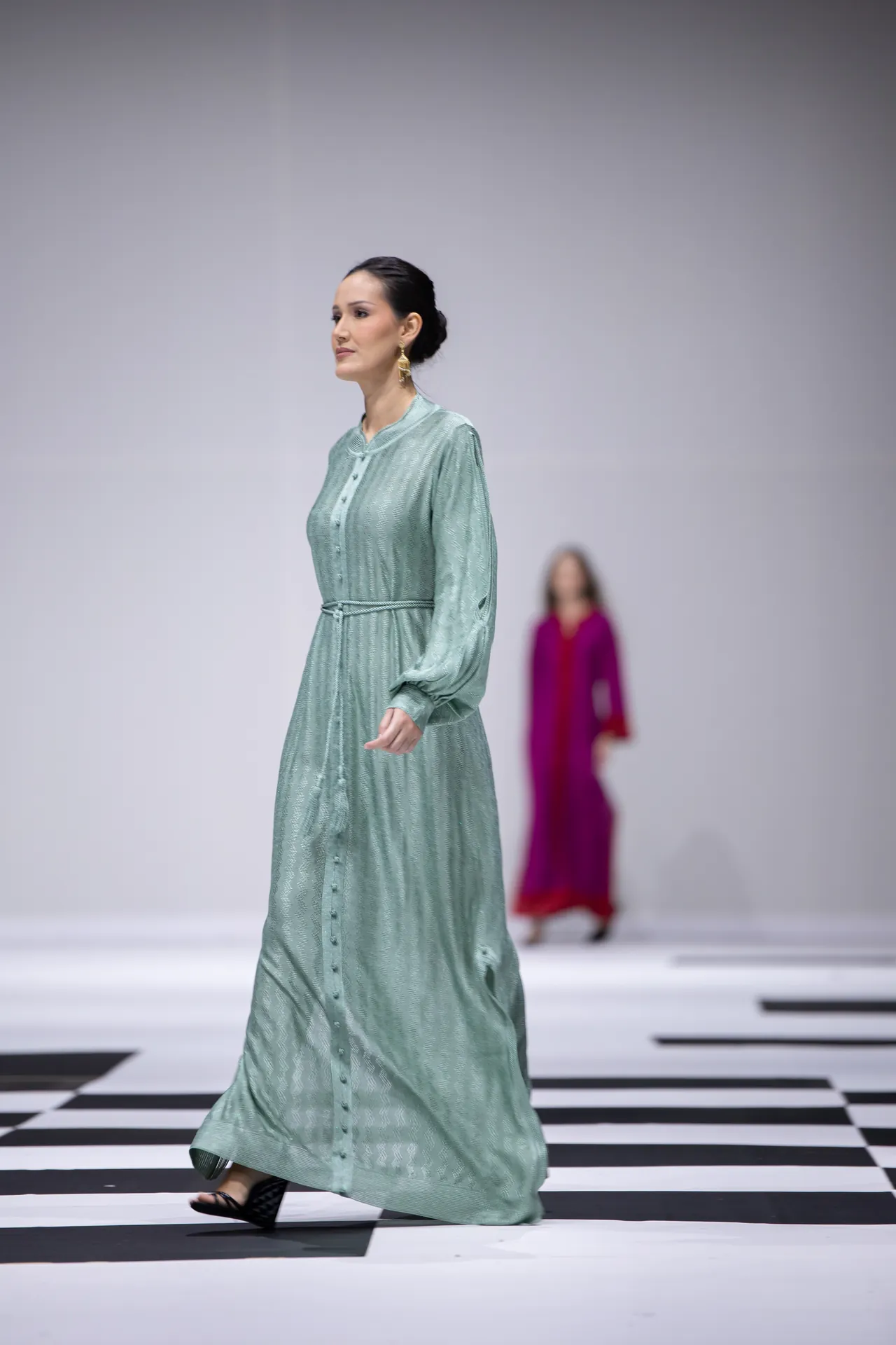 Model walking a mint-green, textured, floor-length dress on a black-and-white runway