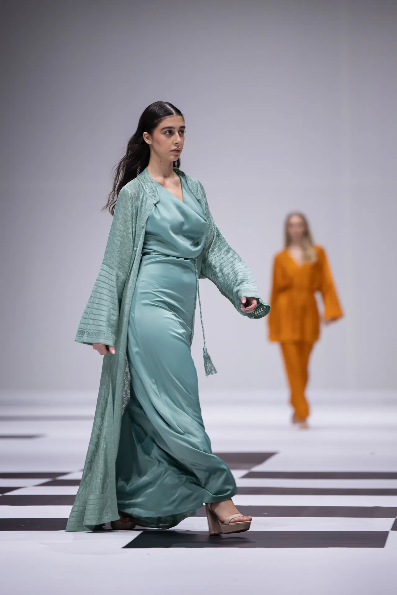 Model walks a turquoise gown and matching cardigan on a black-and-white checkered runway, another orange-outfit model blurred in background