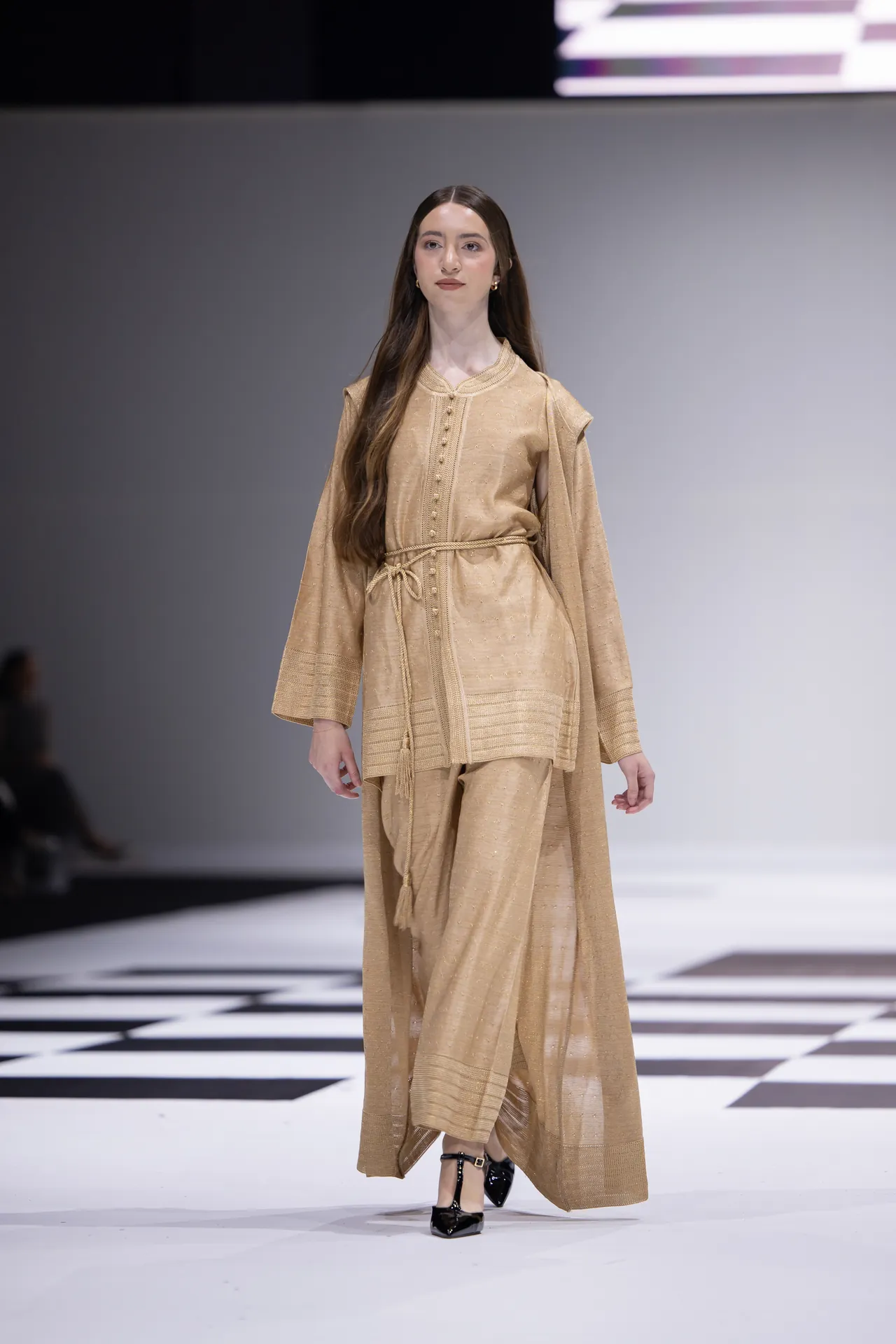 Model walking a fashion show runway in a beige embroidered kaftan with a matching belt and black heels