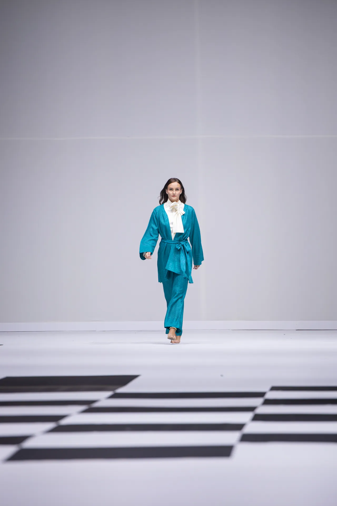 Model walking down a white fashion runway in a teal suit with a white blouse and bow tie.