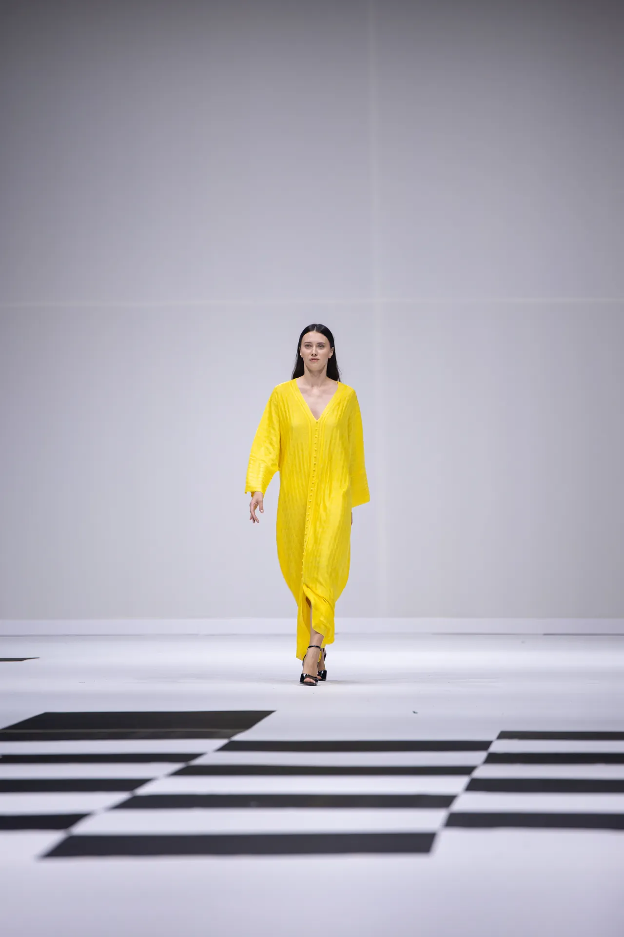 Model walking a minimalist runway in a bright yellow, loose-fitting dress with long sleeves and a V-neck. She wears black high-heeled sandals as she strides forward on a black-and-white geometric runway.