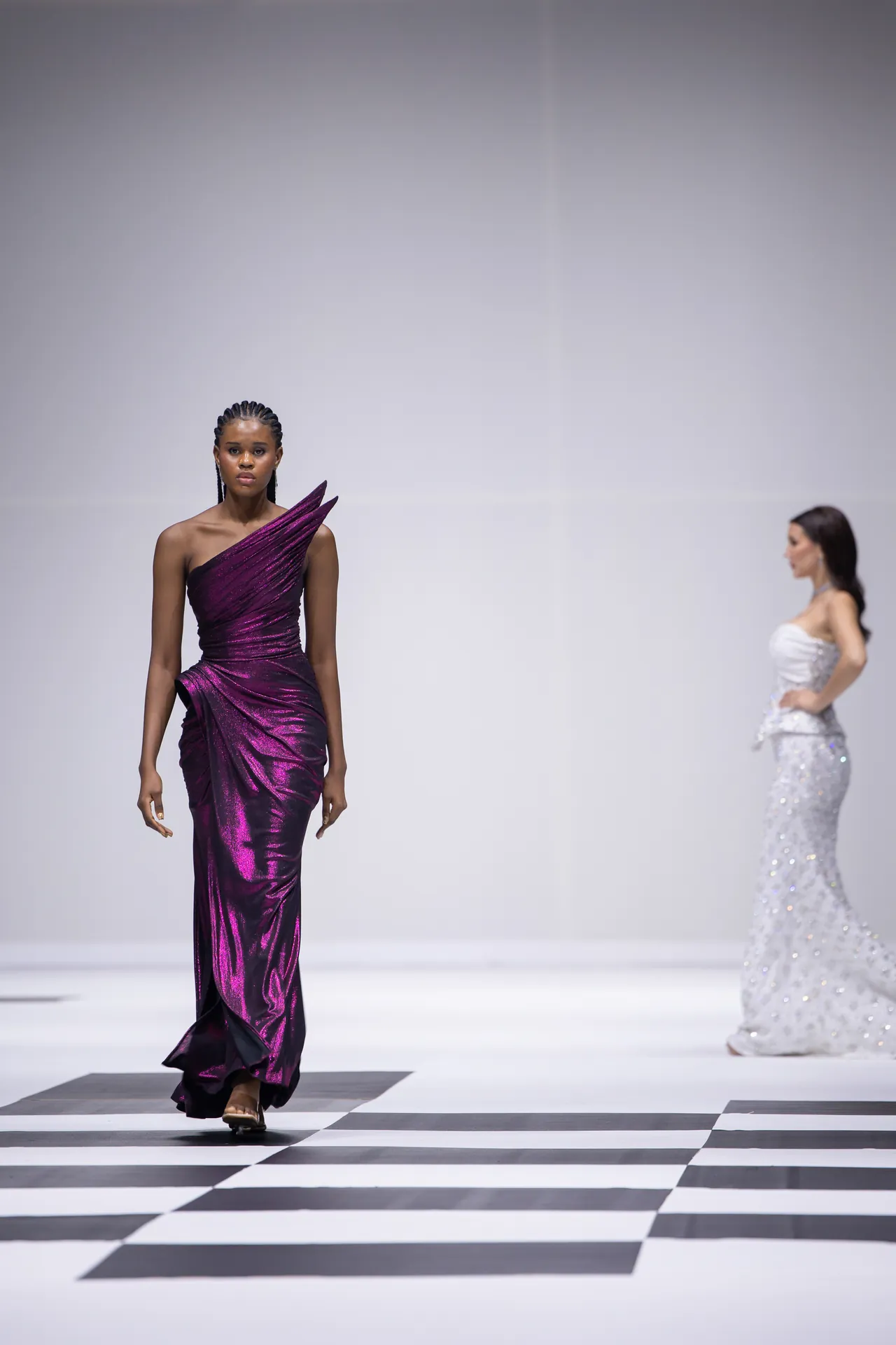 Model in a shiny purple one-shoulder gown walks the runway, with a second model in a white sequined dress in the background on a black-and-white checkered floor.