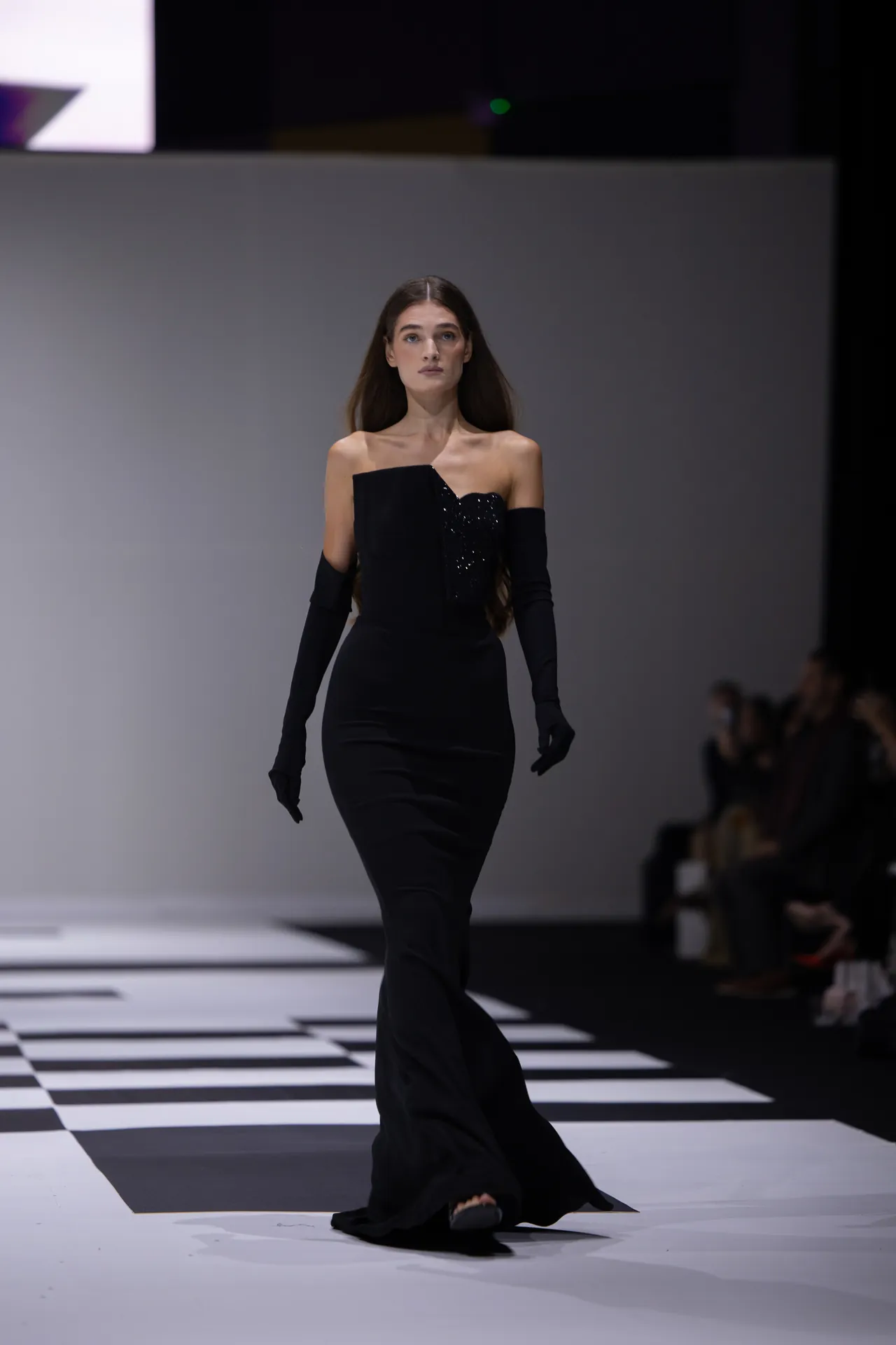 Female model walking a fashion runway in a long black off-the-shoulder gown with gloves, on a black-and-white checkerboard runway.