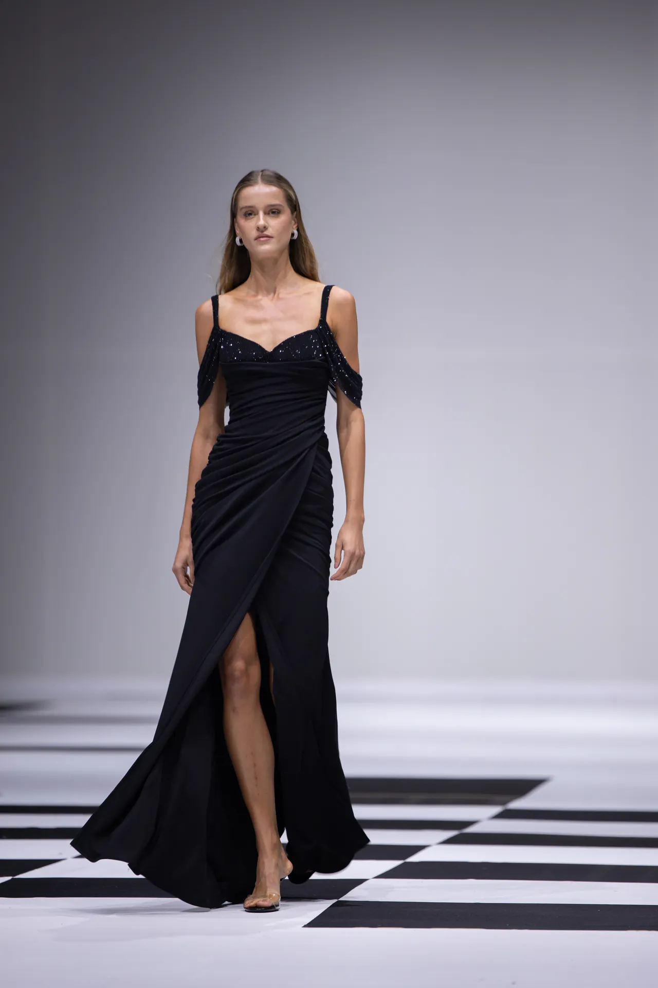 Model walking the runway in a black, off-shoulder gown with draped sleeves on a black-and-white checkered floor seting a fashion show backdrop, full-body view.