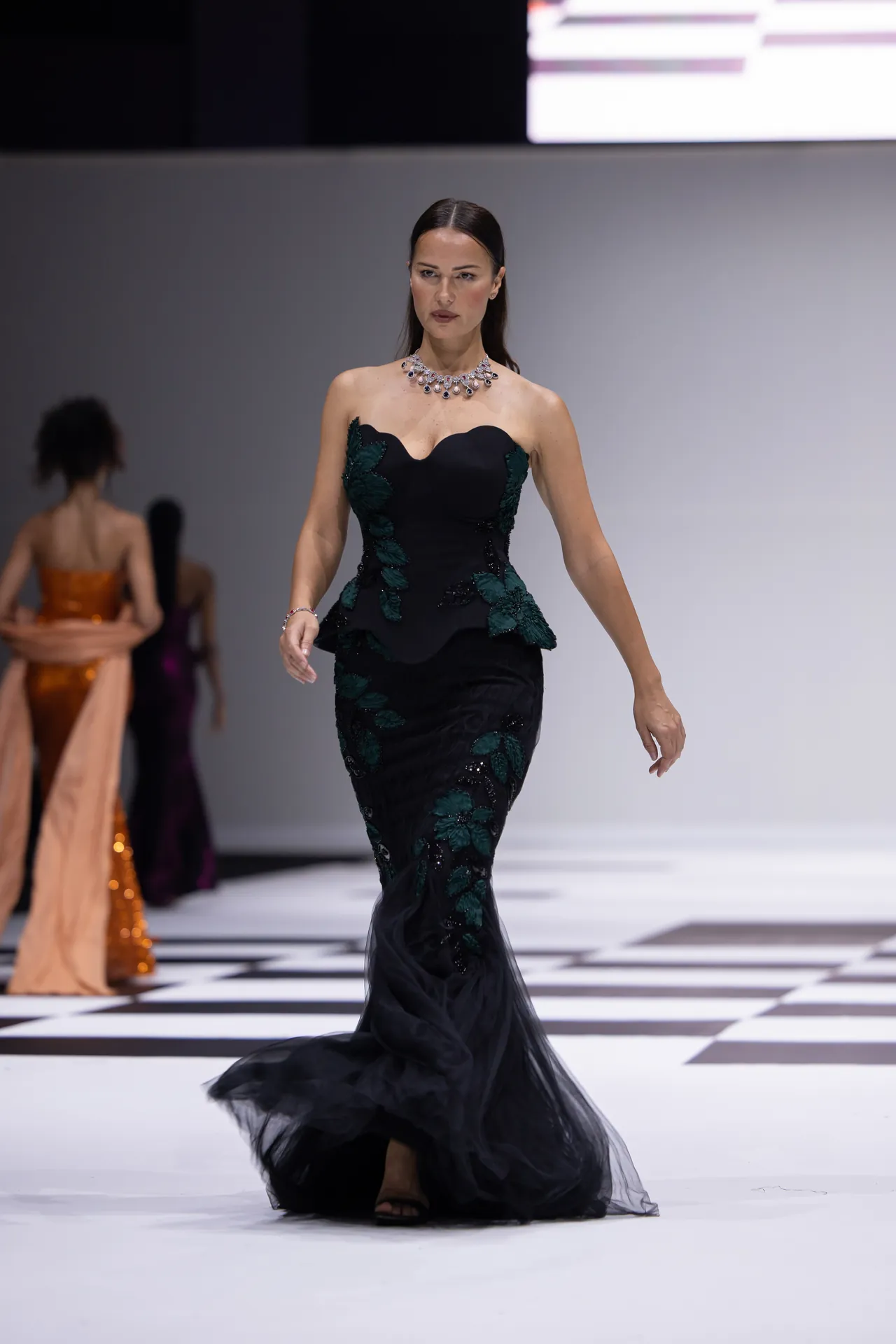 Model walks a runway in a black gown with green leaf embroidery and a statement necklace; checkered floor beneath.