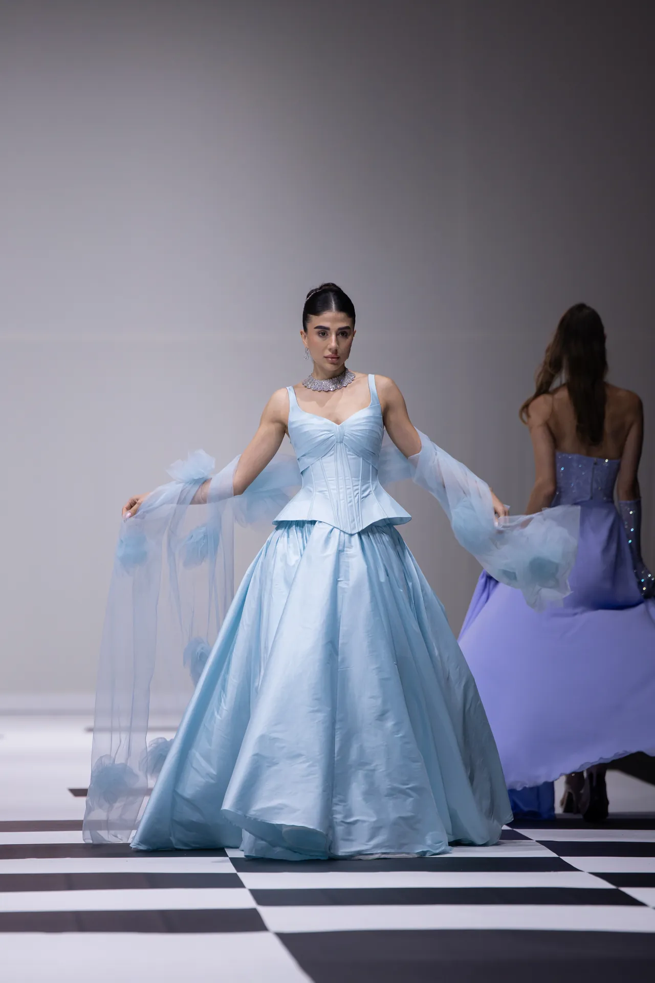 Model in a light blue ball gown with sheer sleeves walking a black-and-white checkerboard runway; another model in blue in the background.
