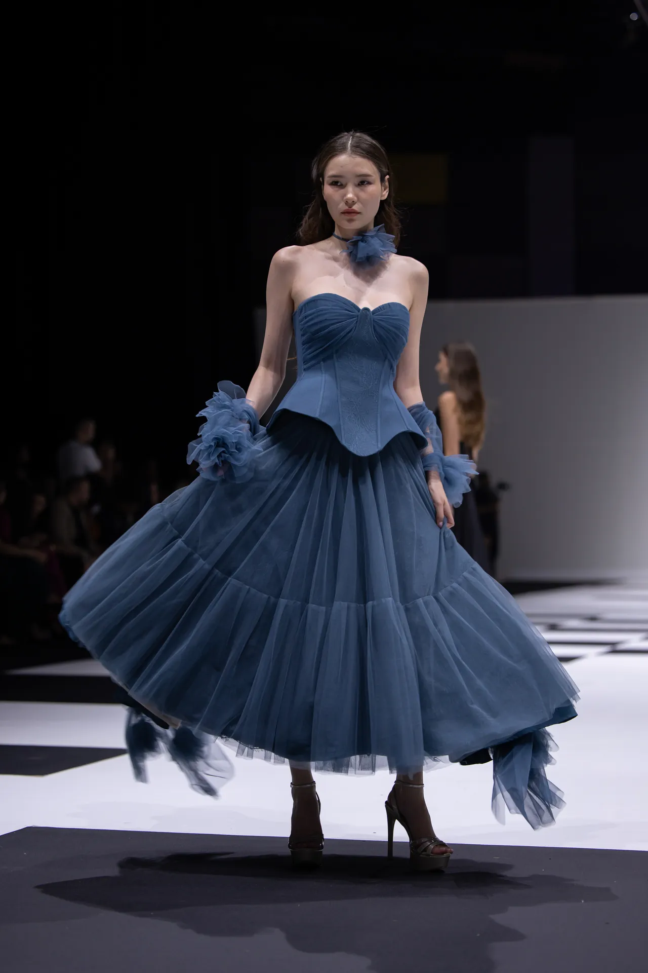 Model on runway wearing a blue strapless ball gown with layered tulle skirt and matching tulle ruffles at wrists, posing under spotlight.