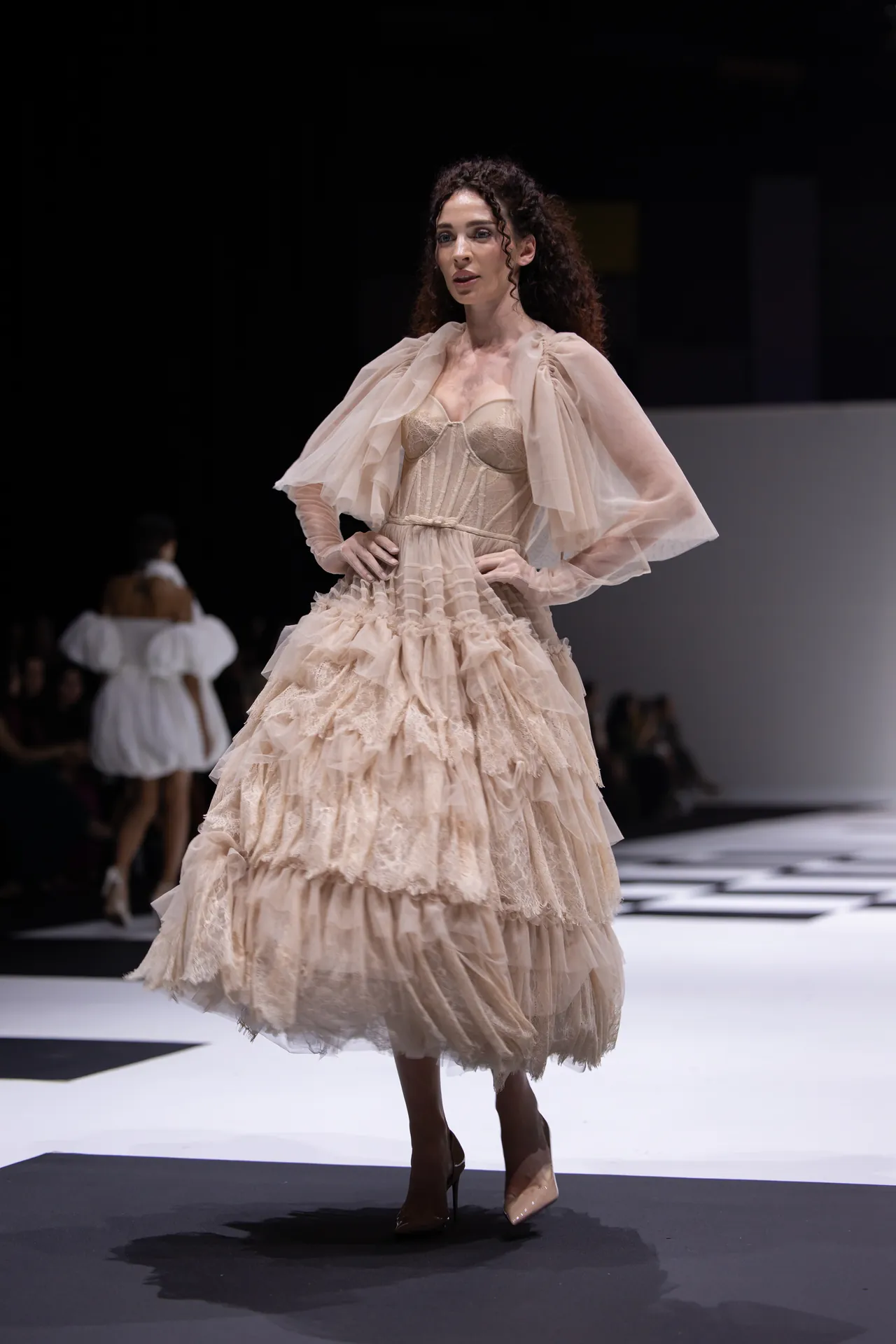 Model walking a fashion show in a voluminous beige lace gown with sheer cape sleeves and ruffled skirt