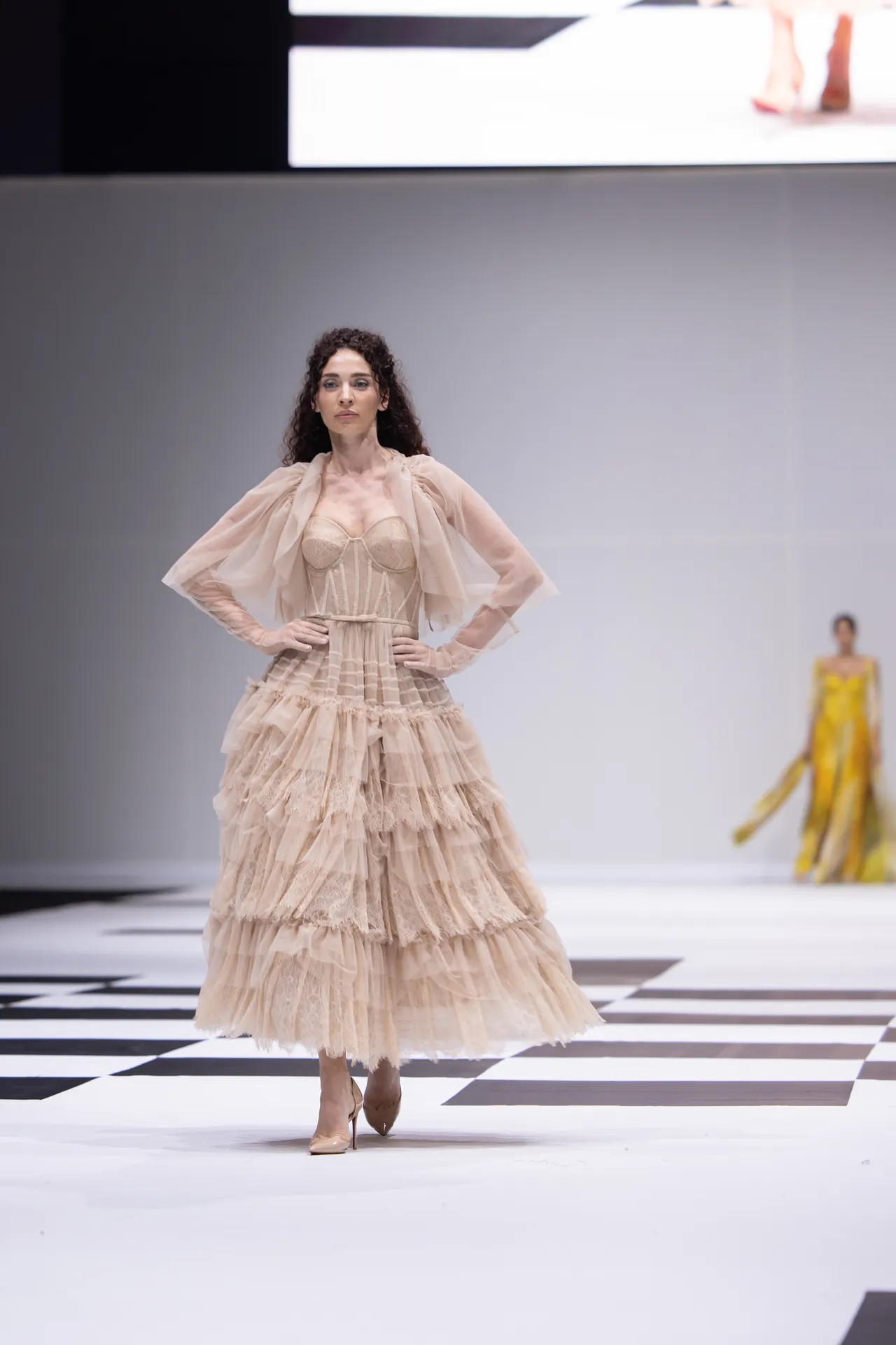 Model walks a fashion runway in a long nude-tone layered gown with sheer sleeves; black-and-white checkered floor and a distant yellow-clad model in background.