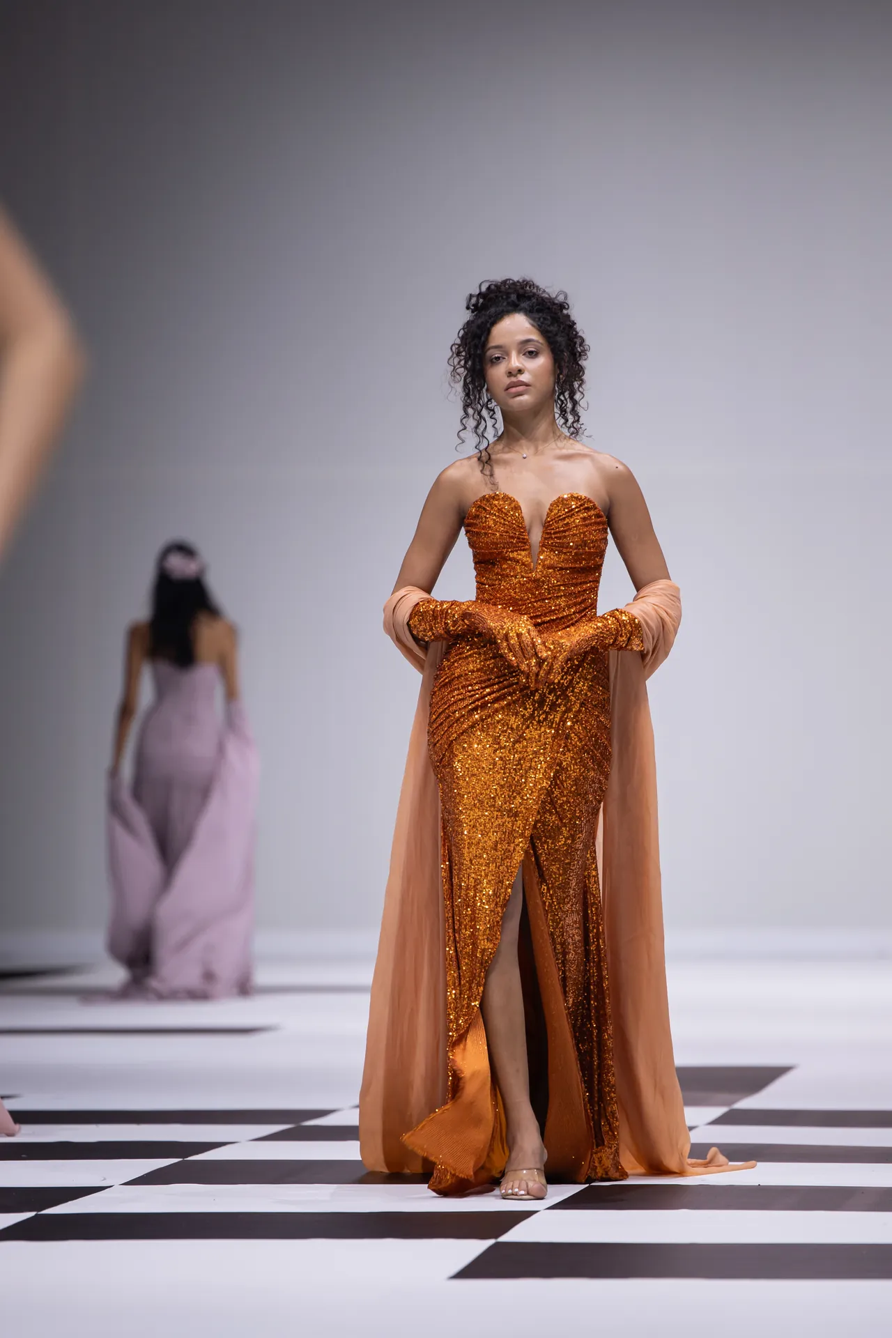 Female model on a fashion runway wearing an orange sequined strapless gown with a peach cape, standing on a black-and-white checkerboard floor.