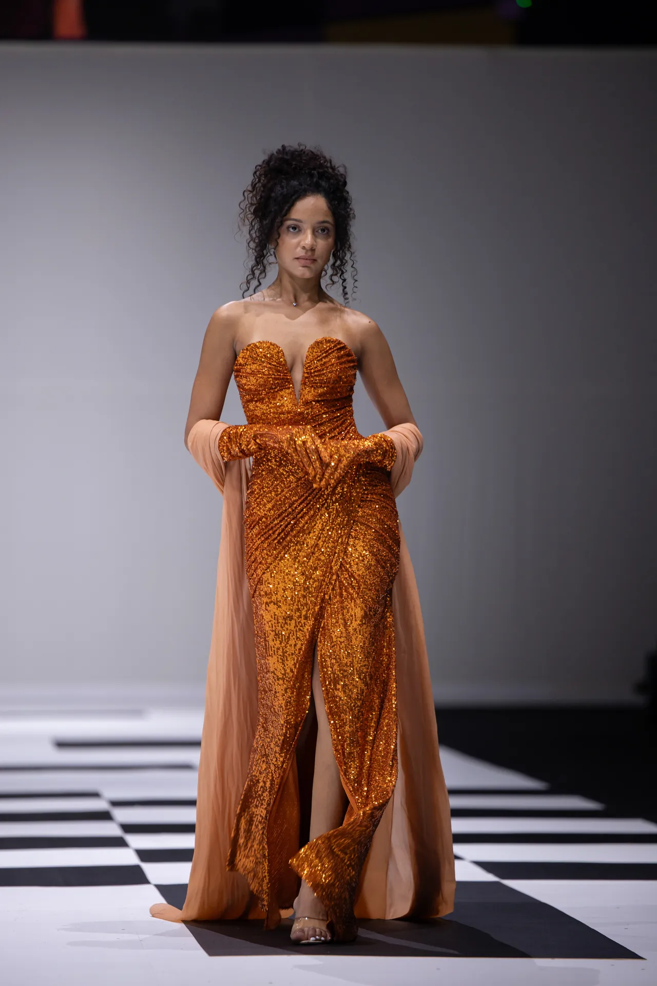 Model wearing orange sequin gown with plunging neckline and peach cape on a black-and-white checker runway.