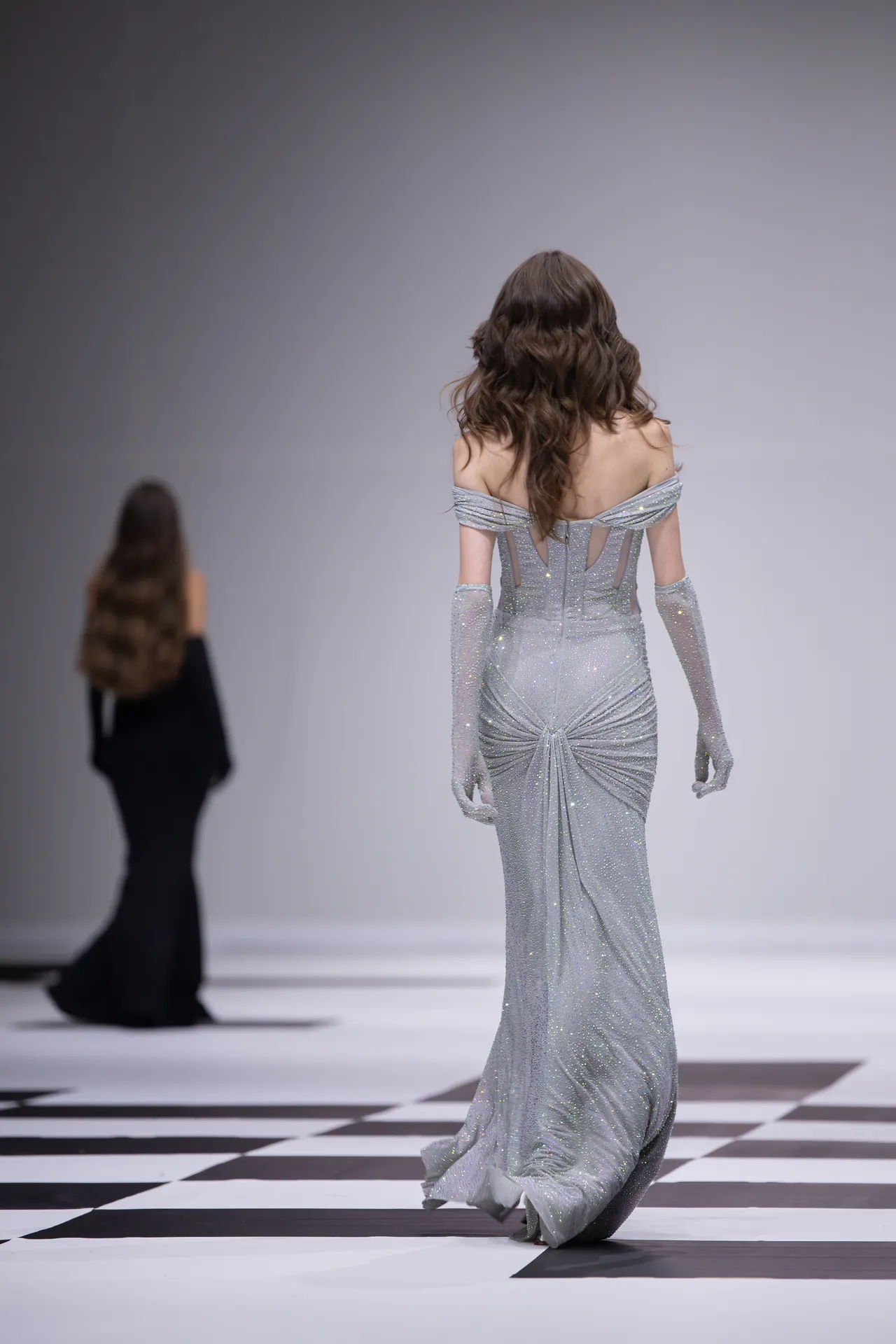 Model walking away on a black-and-white checkered runway in a sparkling silver off-the-shoulder gown with gloves; another model in black is blurred in the background.