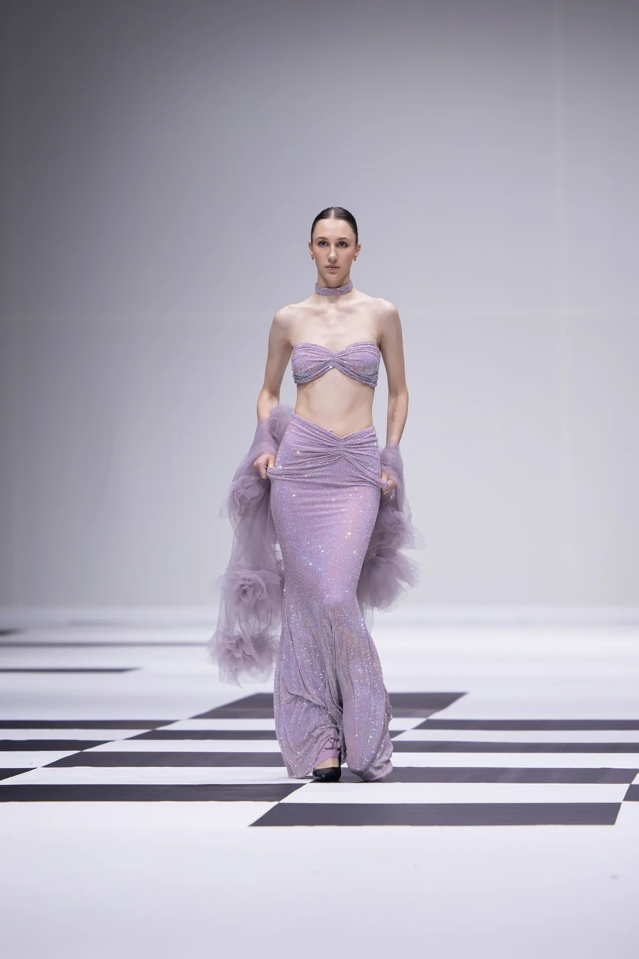 Model walks a lavender sequin two-piece gown with a sparkling strapless top and long matching skirt, on a black-and-white checkered runway.