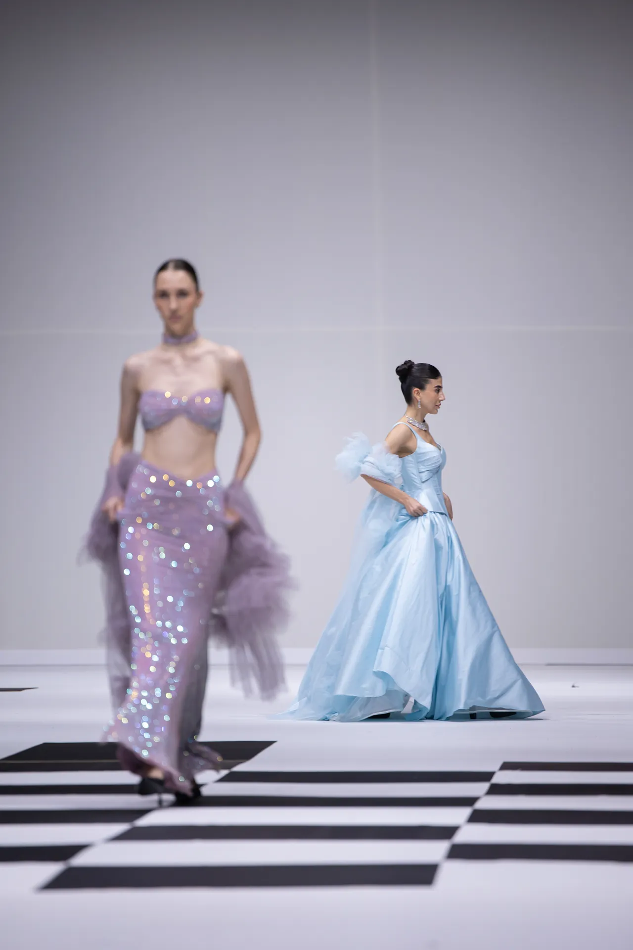 Two models walk a fashion runway; one in a lavender sequin two‑piece with tulle sleeves, the other in a light blue satin ball gown on a black‑and‑white checkerboard catwalk.