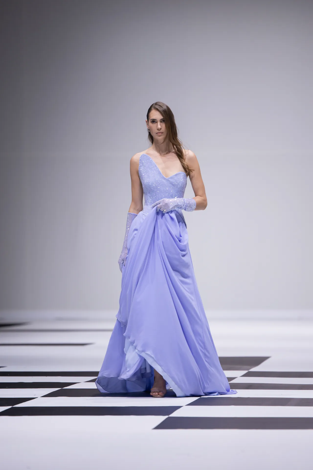 Model walks a lavender, sparkling gown on a black-and-white checkered runway, mid-stride at a fashion show.