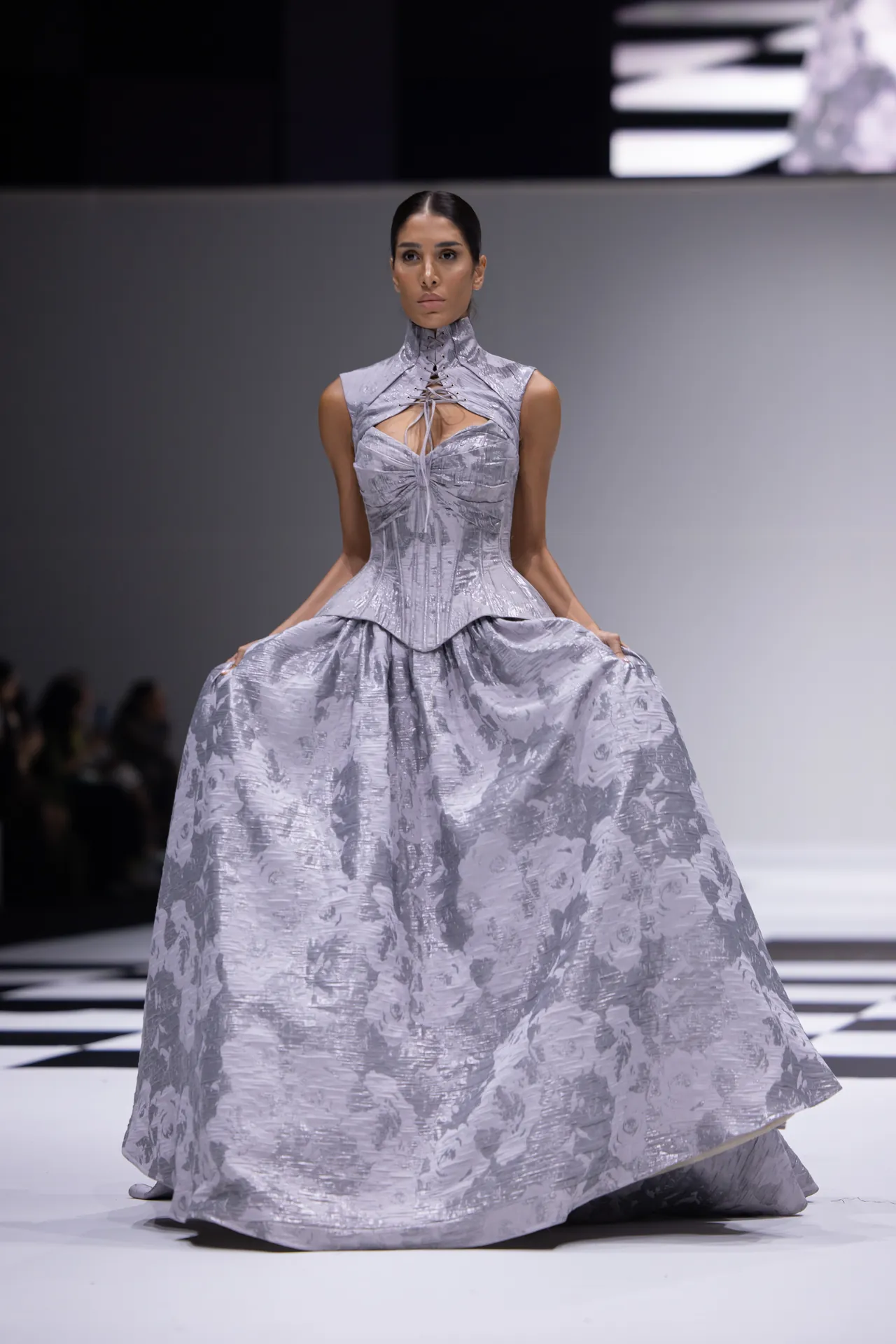 Model on a fashion runway wearing a silver metallic ball gown with a high-neck, corseted bodice and full skirt.