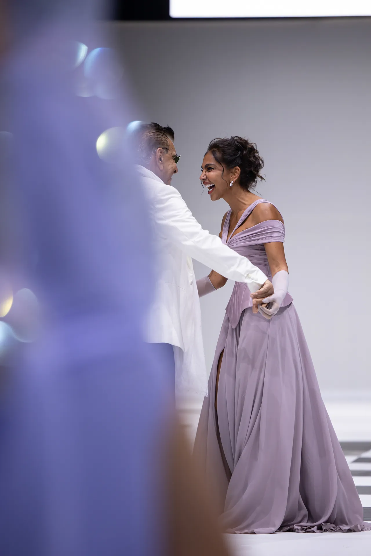 Elegant couple dancing gracefully; woman in lavender evening gown and gloves, man in white suit smiling at her against a light backdrop