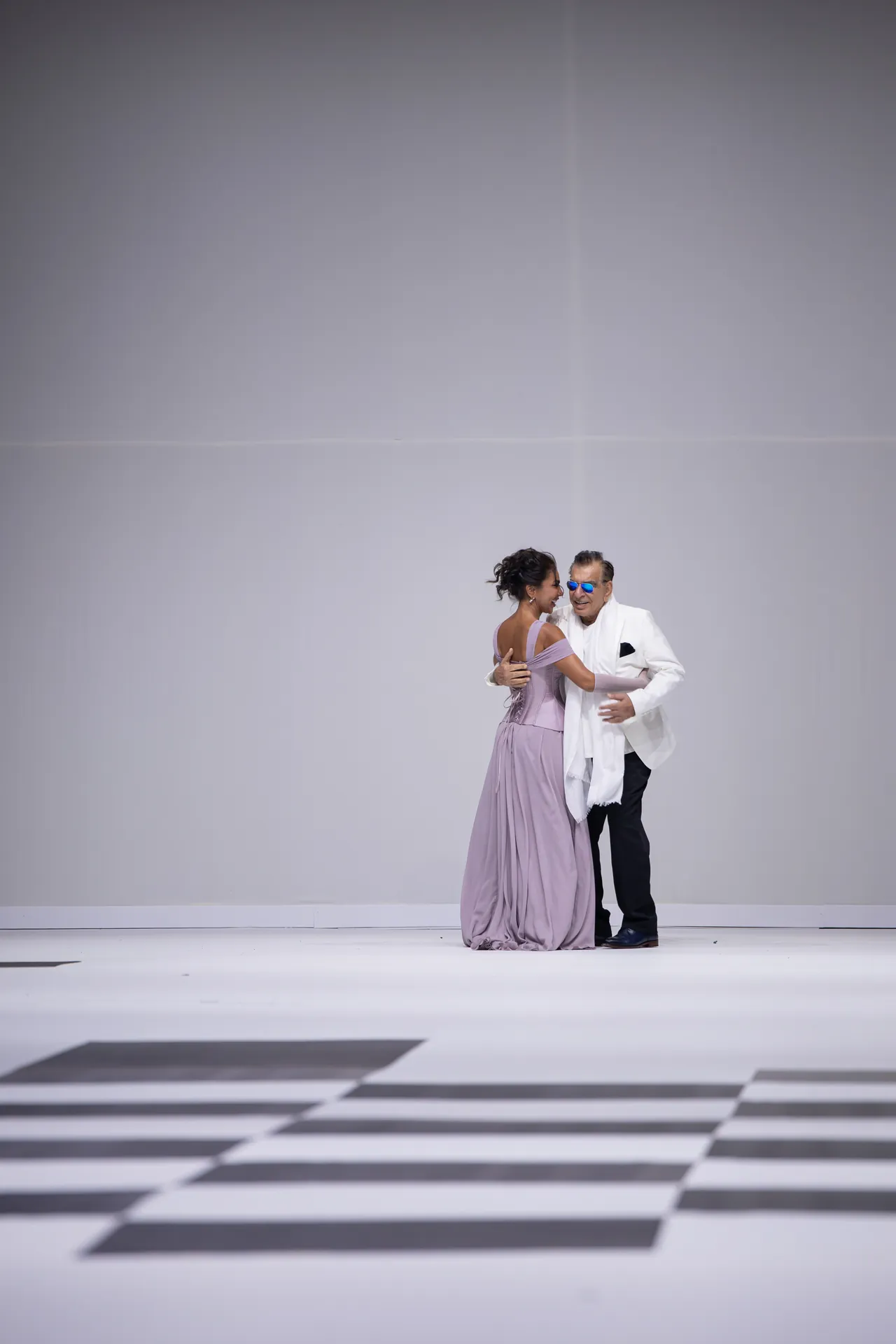 Couple dancing on a white stage; woman in a lavender gown and man in a white suit with blue sunglasses, against a pale backdrop.