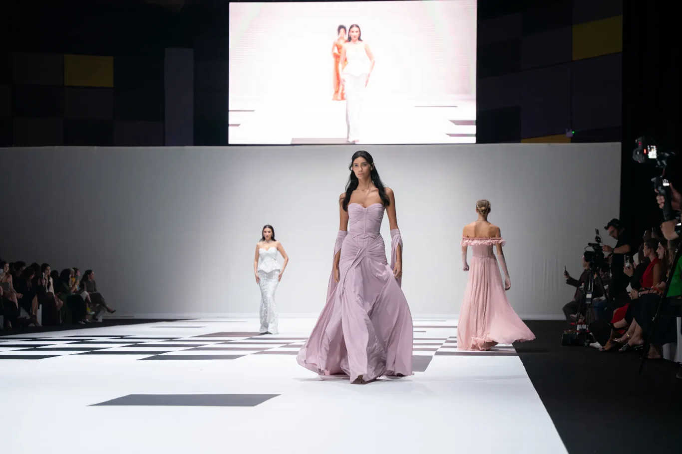 Model in a pale lavender gown walks center-stage on a white checkered runway, with other models in pastel dresses visible behind and to the sides. Audience and cameras line the sides. A large screen above shows a model walking.