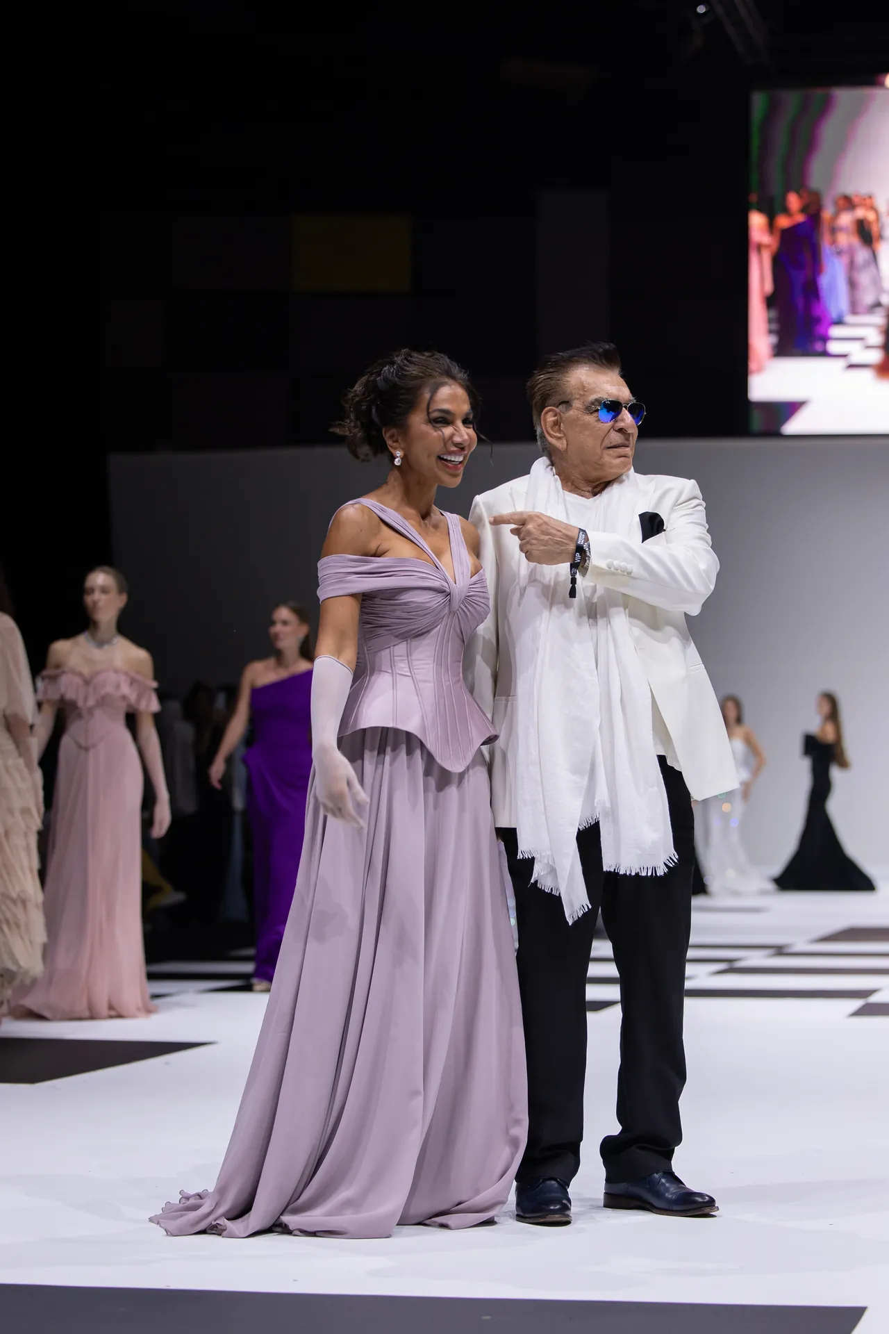 Woman in a lavender off-shoulder gown smiles while walking the runway beside a man in a white suit. Other models are visible in the background.