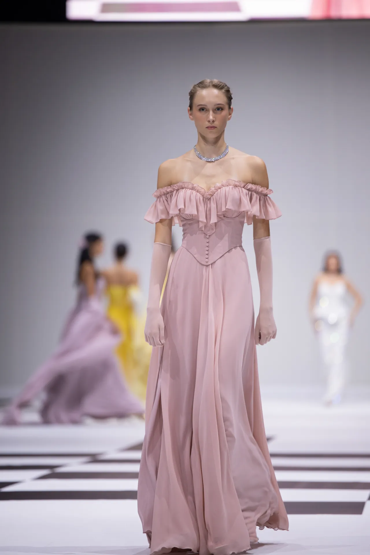 Model in a pastel pink off-the-shoulder gown with ruffled neckline and long gloves walks a fashion run­way, wearing a sparkling necklace.