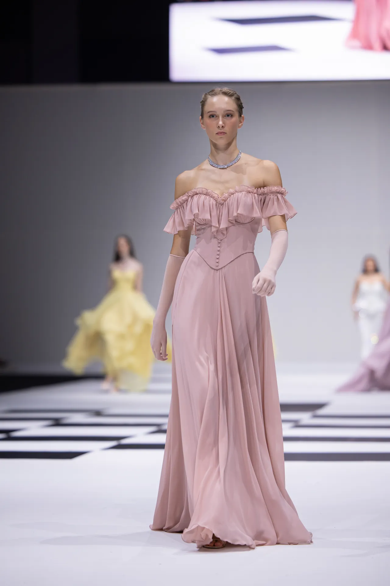 Model walks the runway in a blush pink off-shoulder gown with ruffled neckline and long gloves on a checkerboard catwalk.