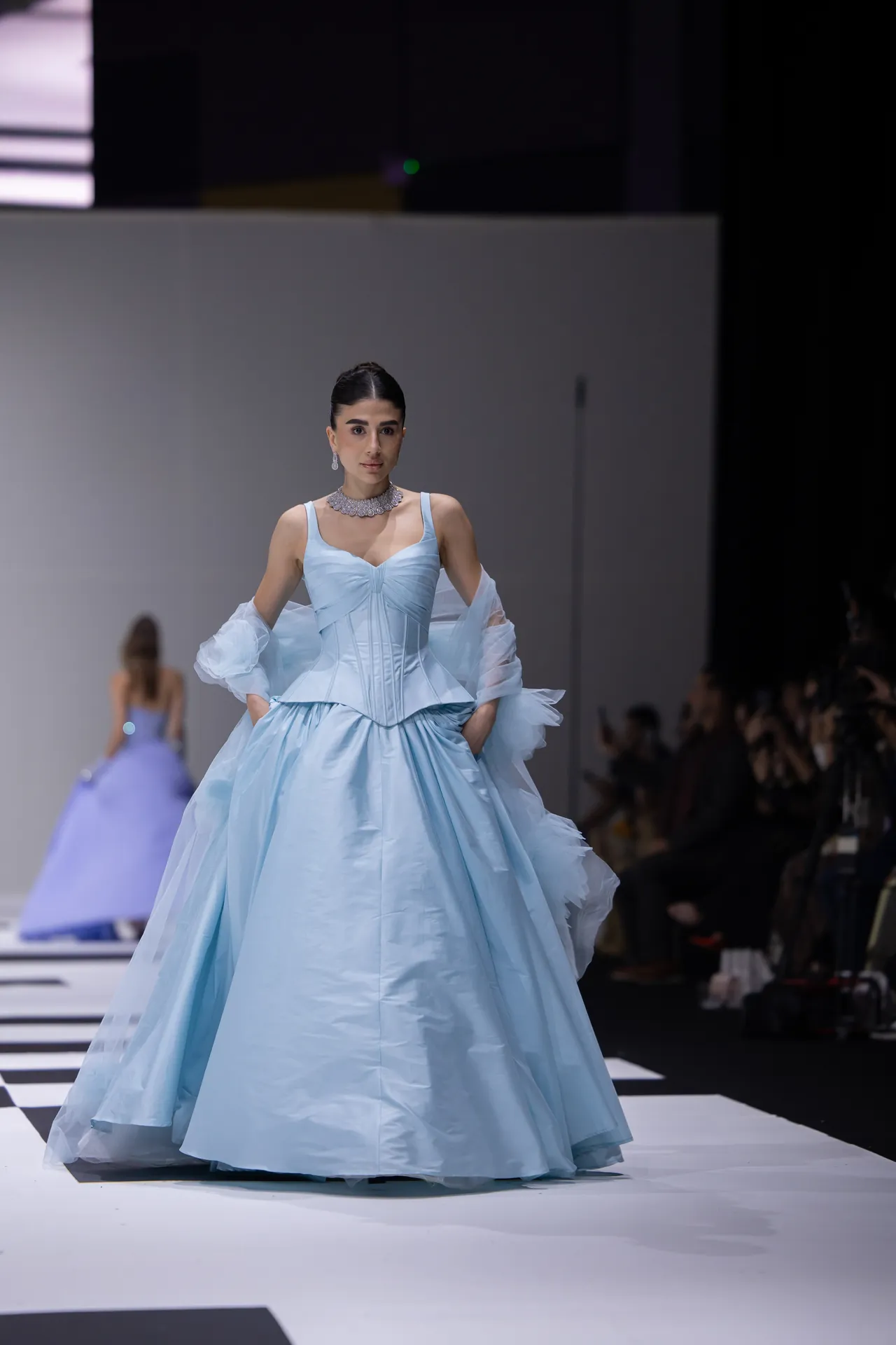 Model walking the runway in a light blue ball gown with sheer, puffed sleeves and a statement necklace.