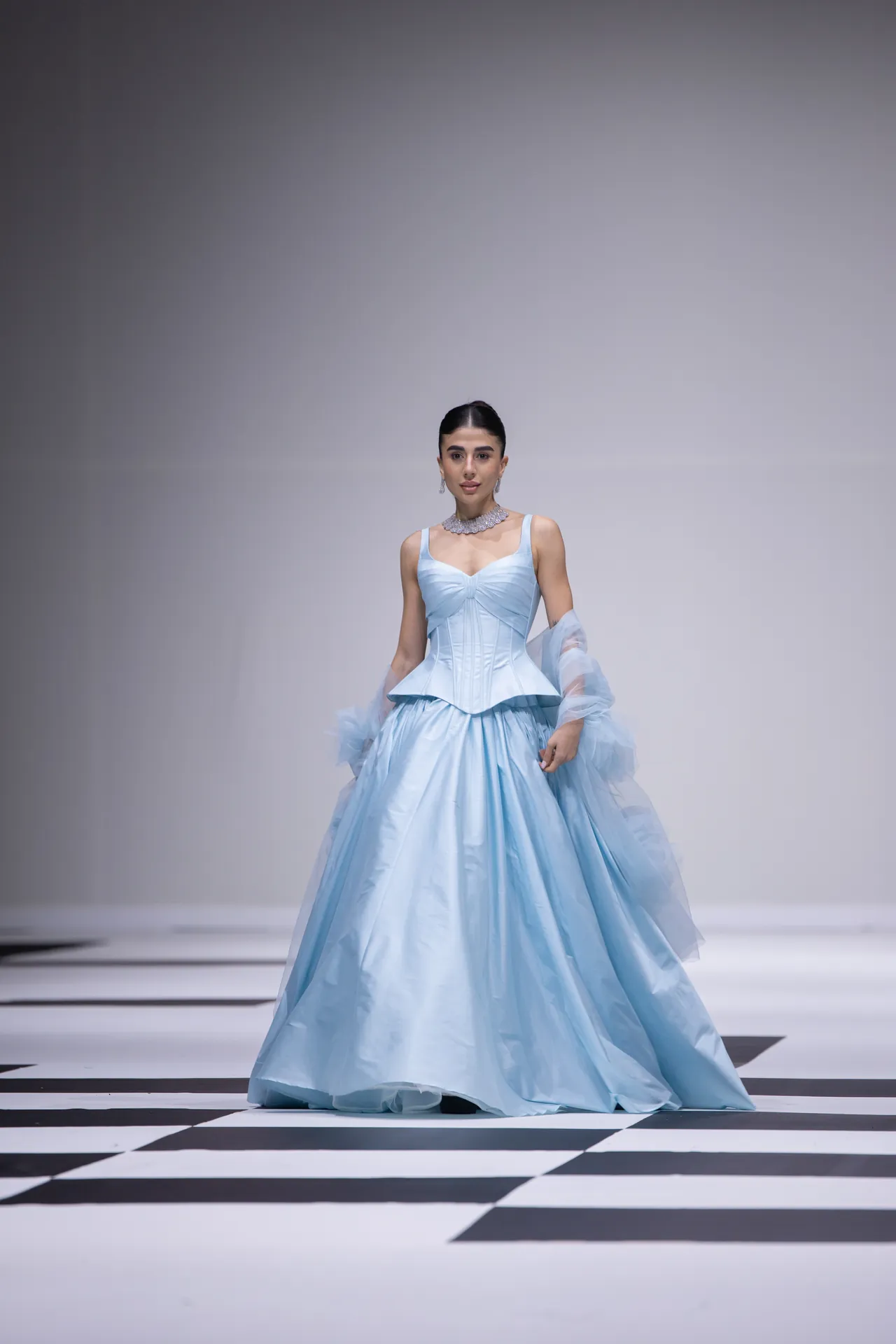 Model on a fashion runway wearing a pale blue, full-skirt ball gown with a fitted corseted bodice and sheer cape sleeves.