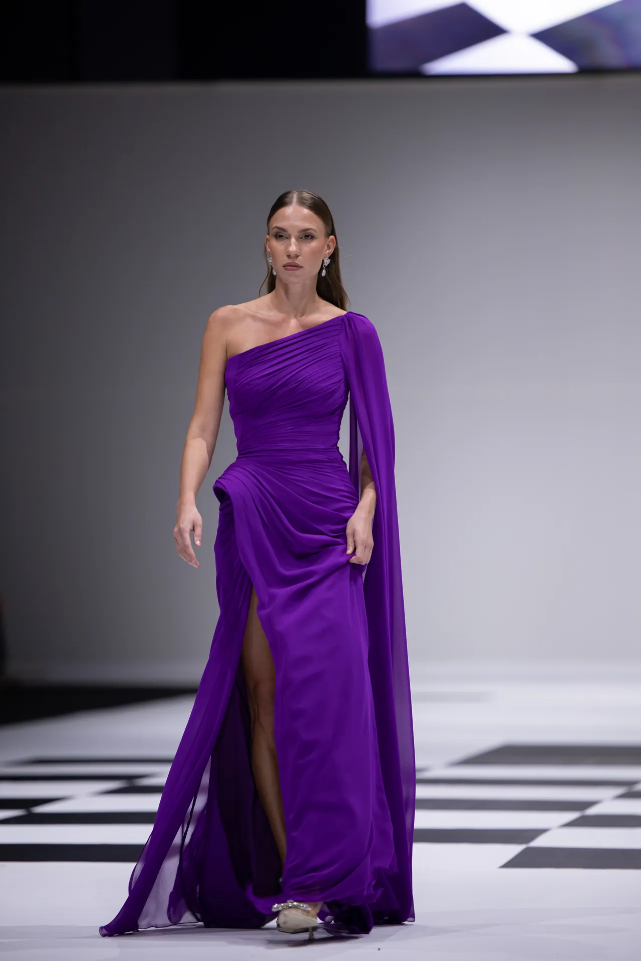 Model walks a runway in a vivid purple, one-shoulder gown with draped fabric and cape-like sleeve on a black-and-white checkered floor.