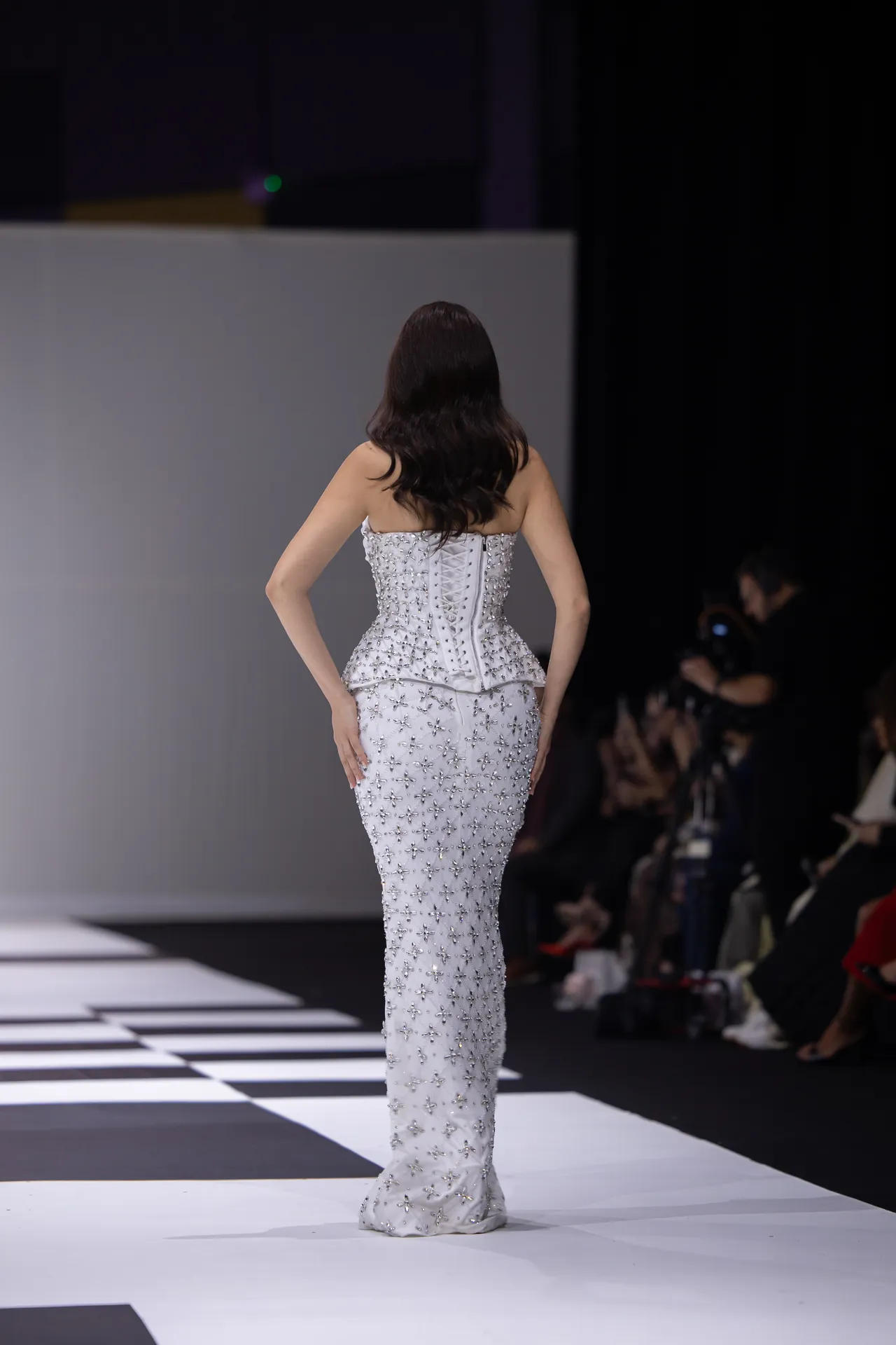 Back view of a model walking the runway in a strapless, silver beaded mermaid gown; audience and photographers line the sides in a dimly lit fashion show setting.