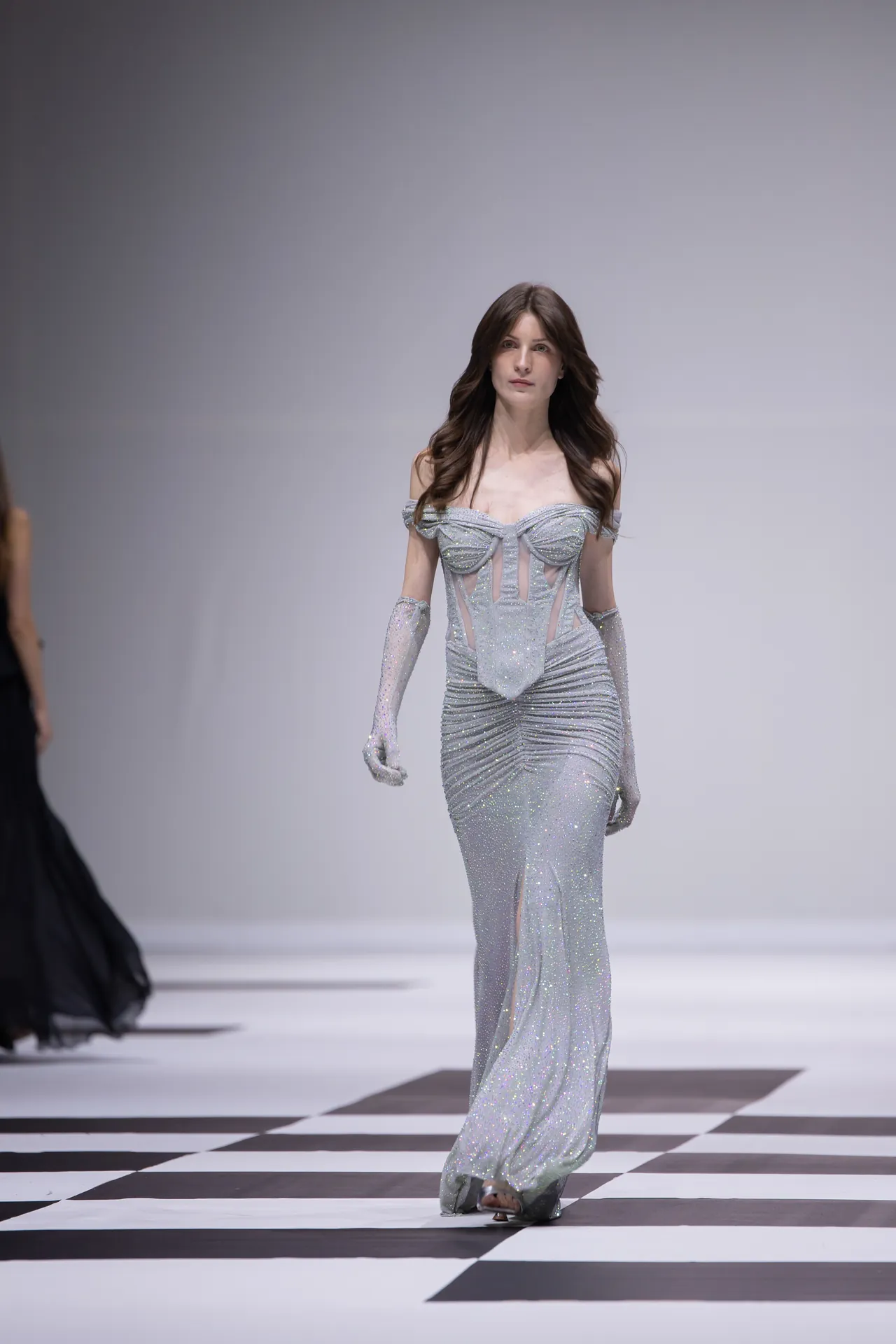 Model walking a checkered runway in a sparkly silver off-shoulder gown with long gloves.