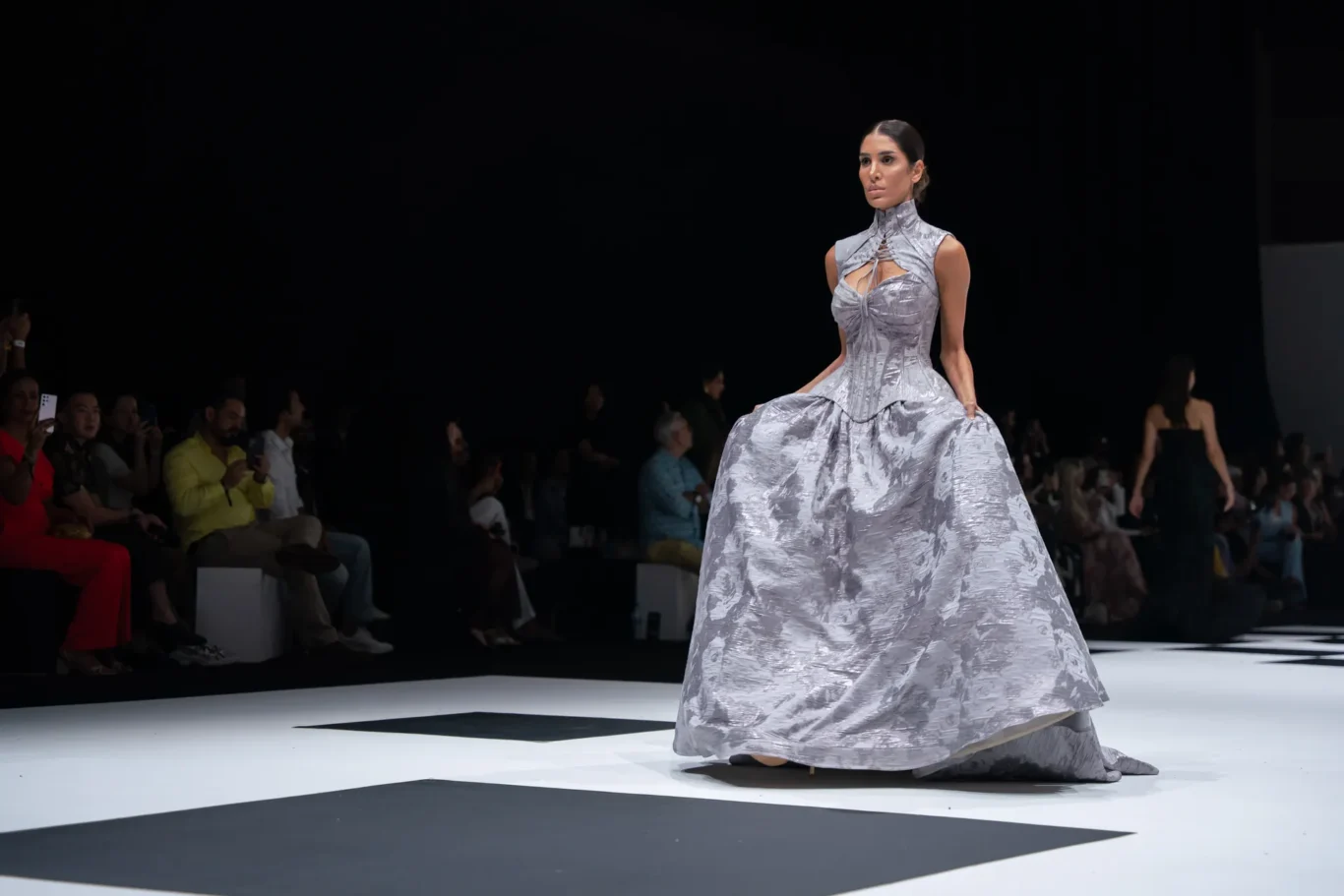 Model walks a voluminous metallic silver gown down the runway as the audience watches from the left.