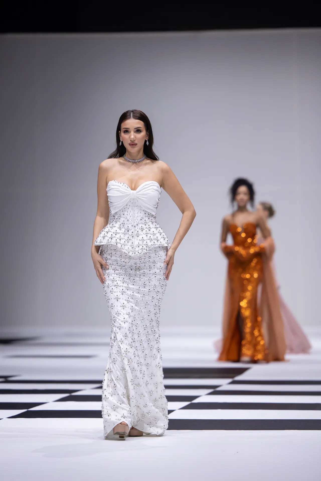 Model wearing a strapless white beaded gown on a runway, with blurred background models behind her.