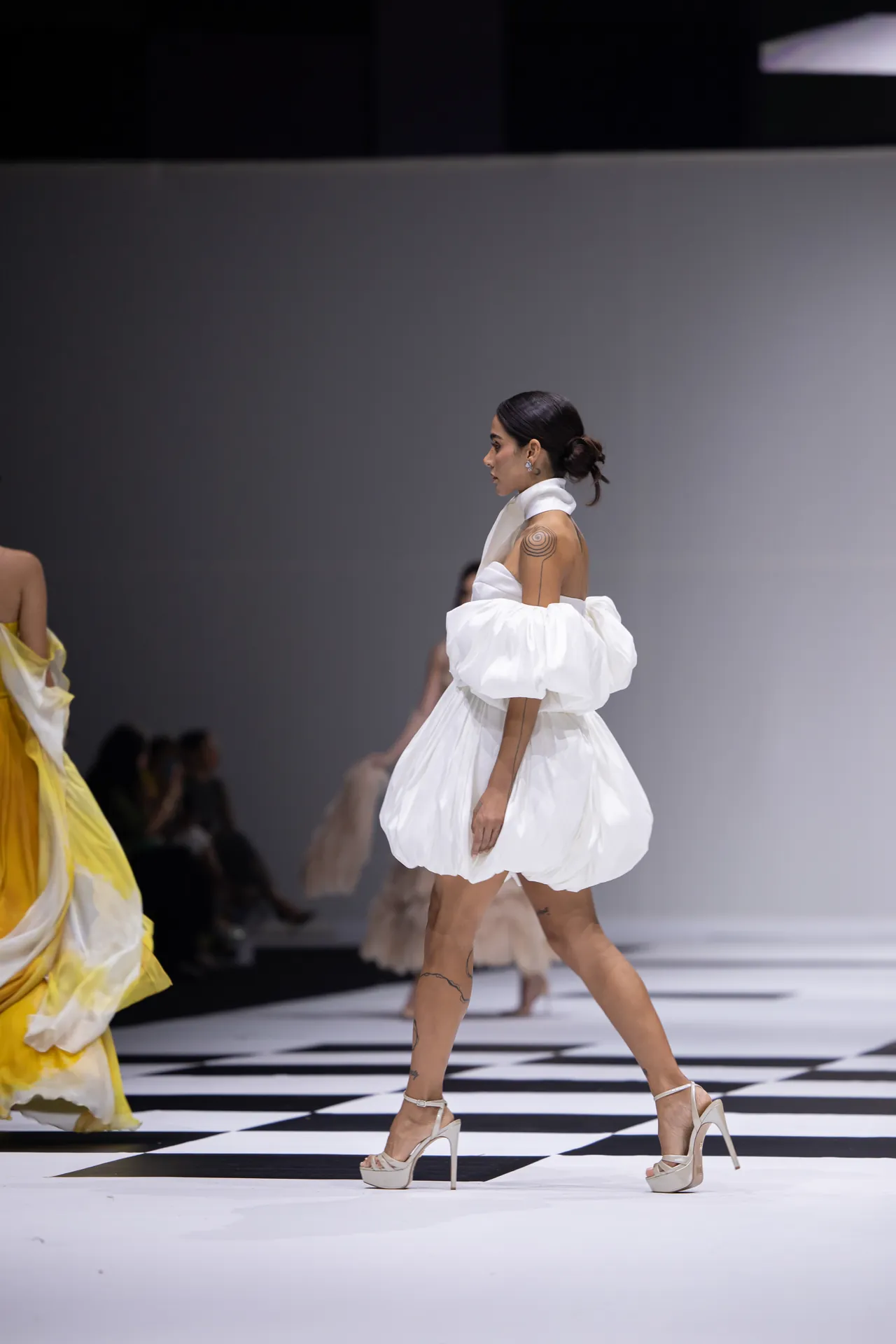 Model walking a fashion show in a white voluminous off-shoulder dress and high-heeled sandals on a black-and-white checkered runway, tattoos visible on her arm.