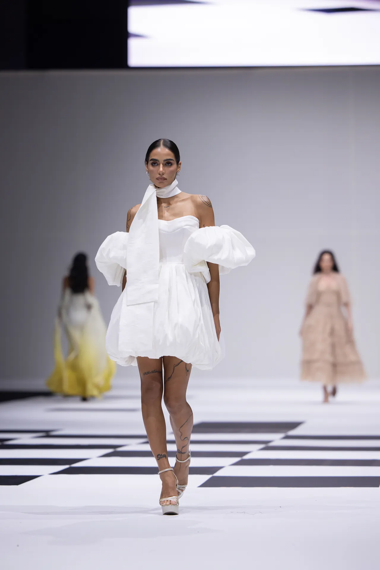 Model walks a white, sculptural dress with oversized puff sleeves down a black-and-white checkerboard runway at a fashion show.
