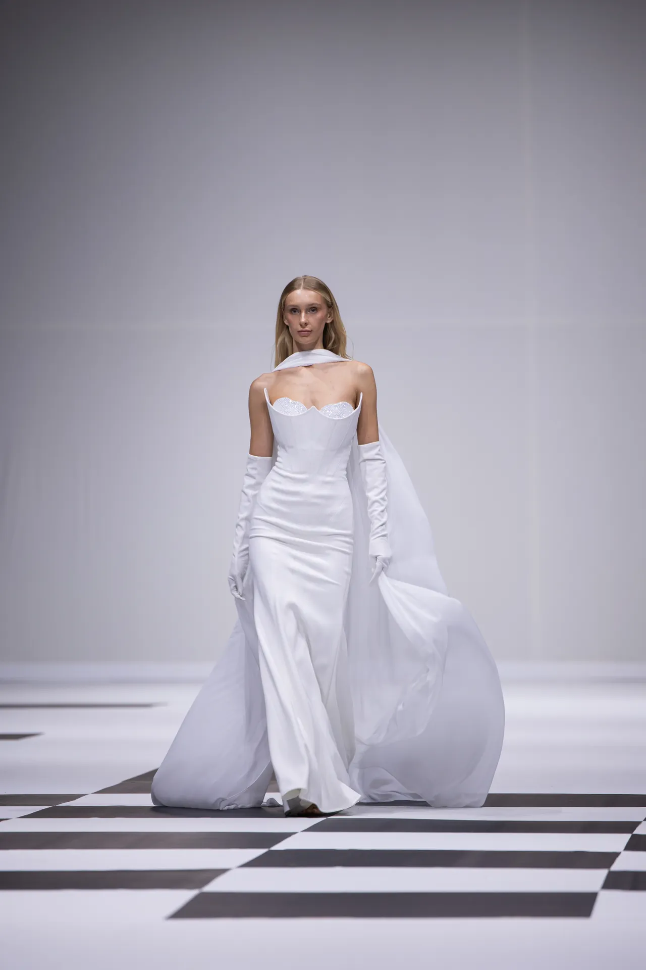 Model walking a sleek white bridal gown with long white gloves on a black-and-white checkerboard runway.