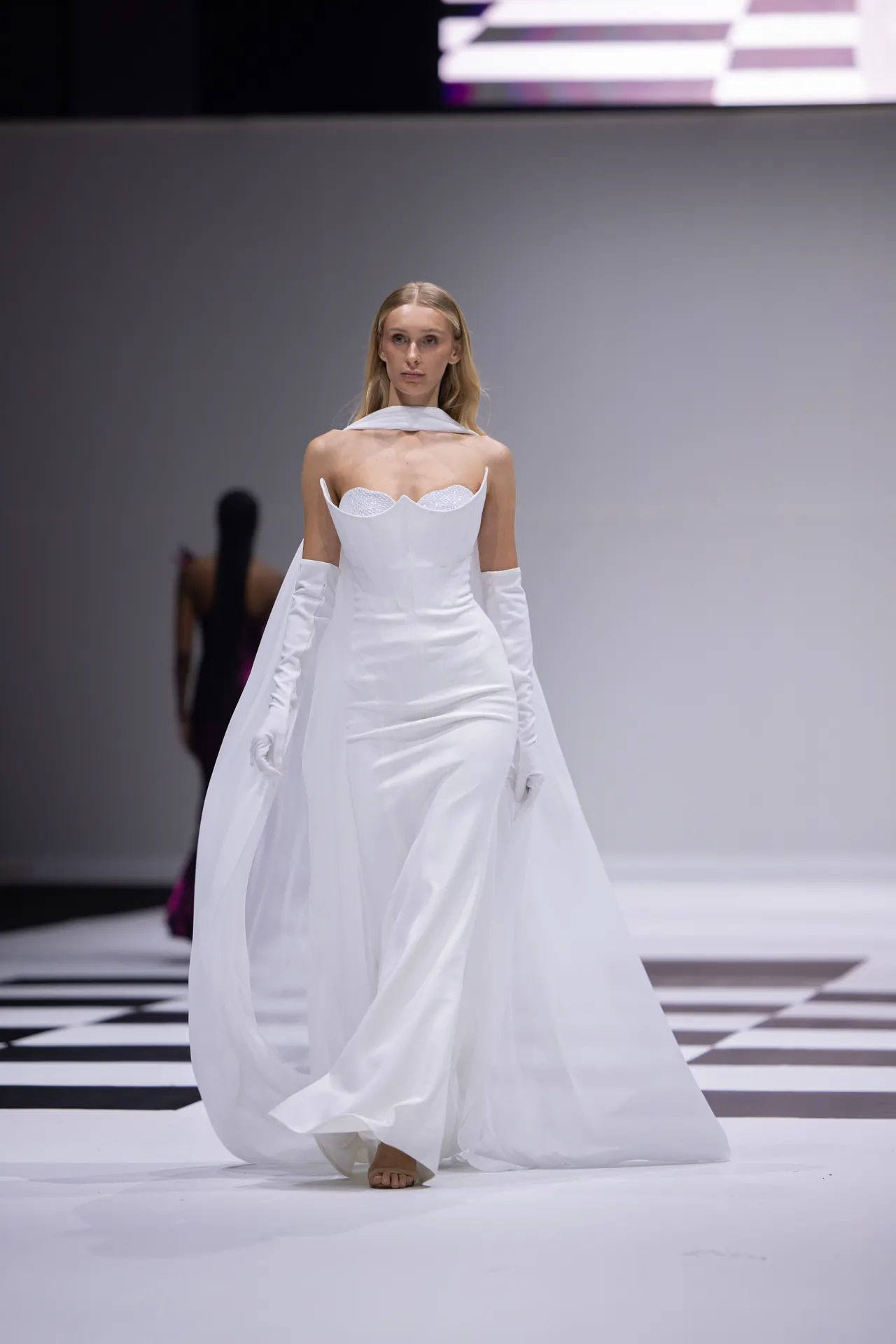 Model walking a fashion runway in a white, structured wedding-style gown with long white gloves and cape, on a black-and-white checkered floor.