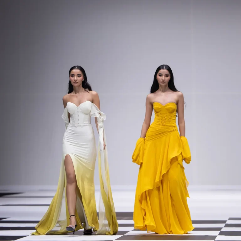 Two female models walk a fashion runway in elegant gowns: one white with off-the-shoulder sleeves and a high slit, the other bright yellow with ruffled layers and gloves-like details on the arms.