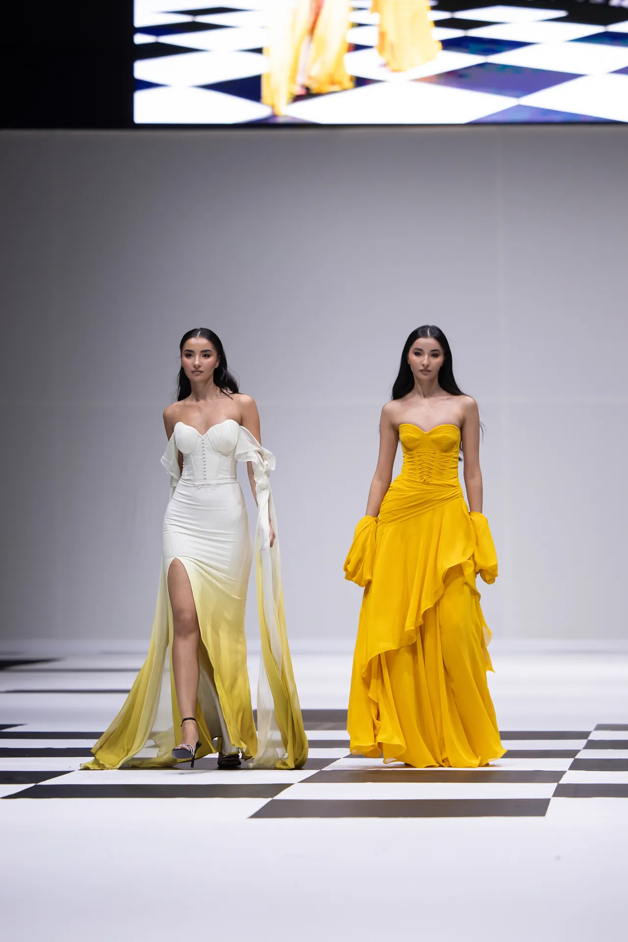 Two female models walk a fashion runway in elegant gowns: one white with off-the-shoulder sleeves and a high slit, the other bright yellow with ruffled layers and gloves-like details on the arms.