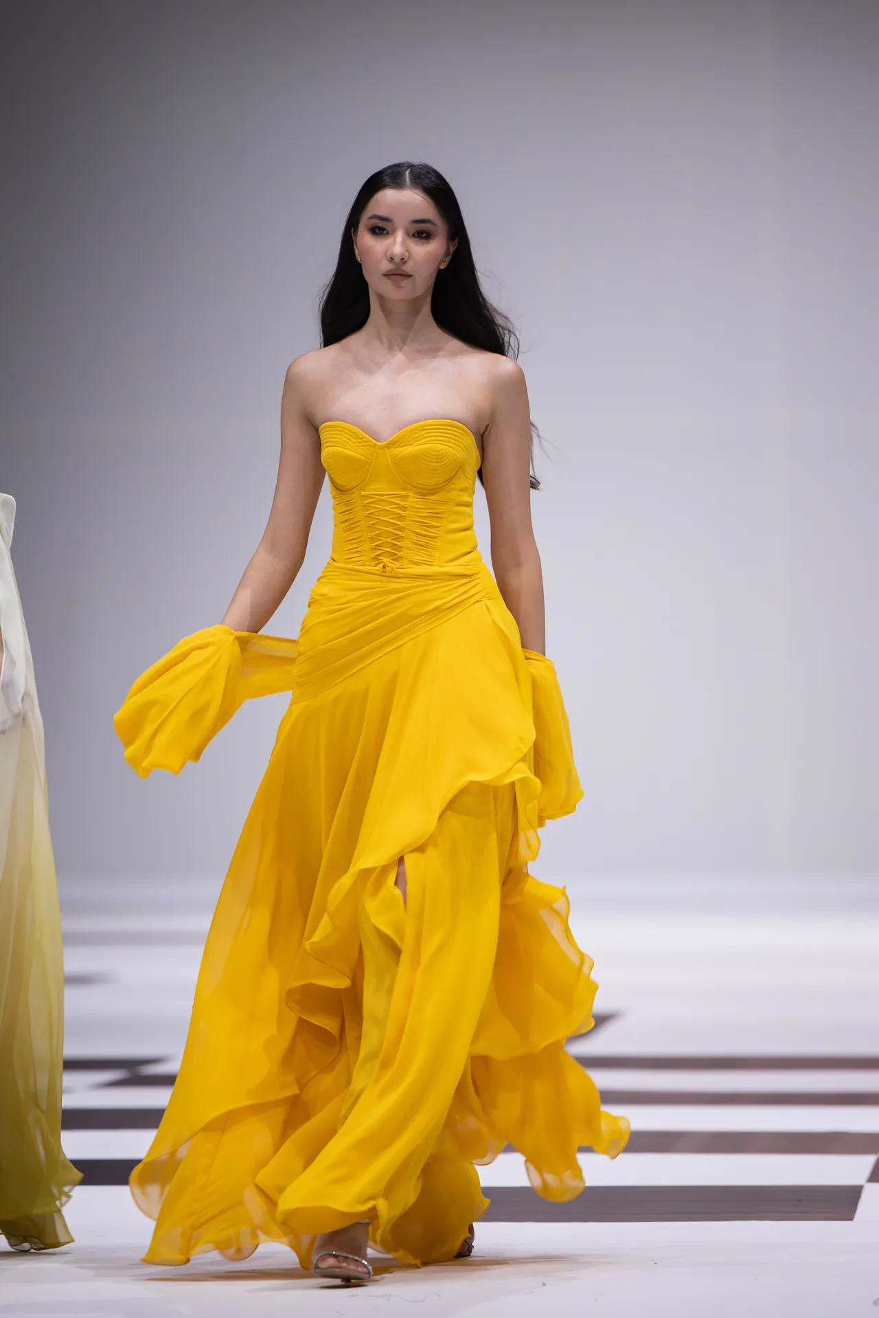 Model walking the runway in a strapless bright yellow gown with a corseted bodice and flowing skirt.