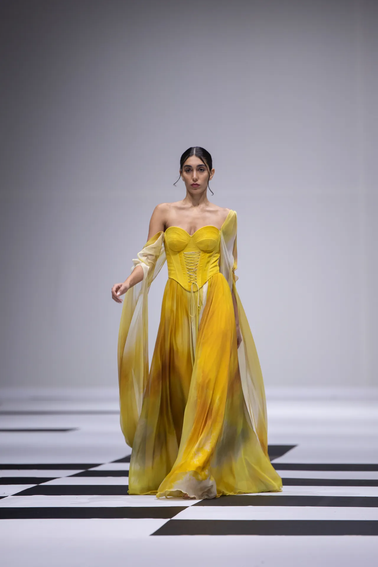 Model walking on a runway in a yellow gradient ball gown with a corset bodice and sheer sleeves, against a black-and-white checkered floor.