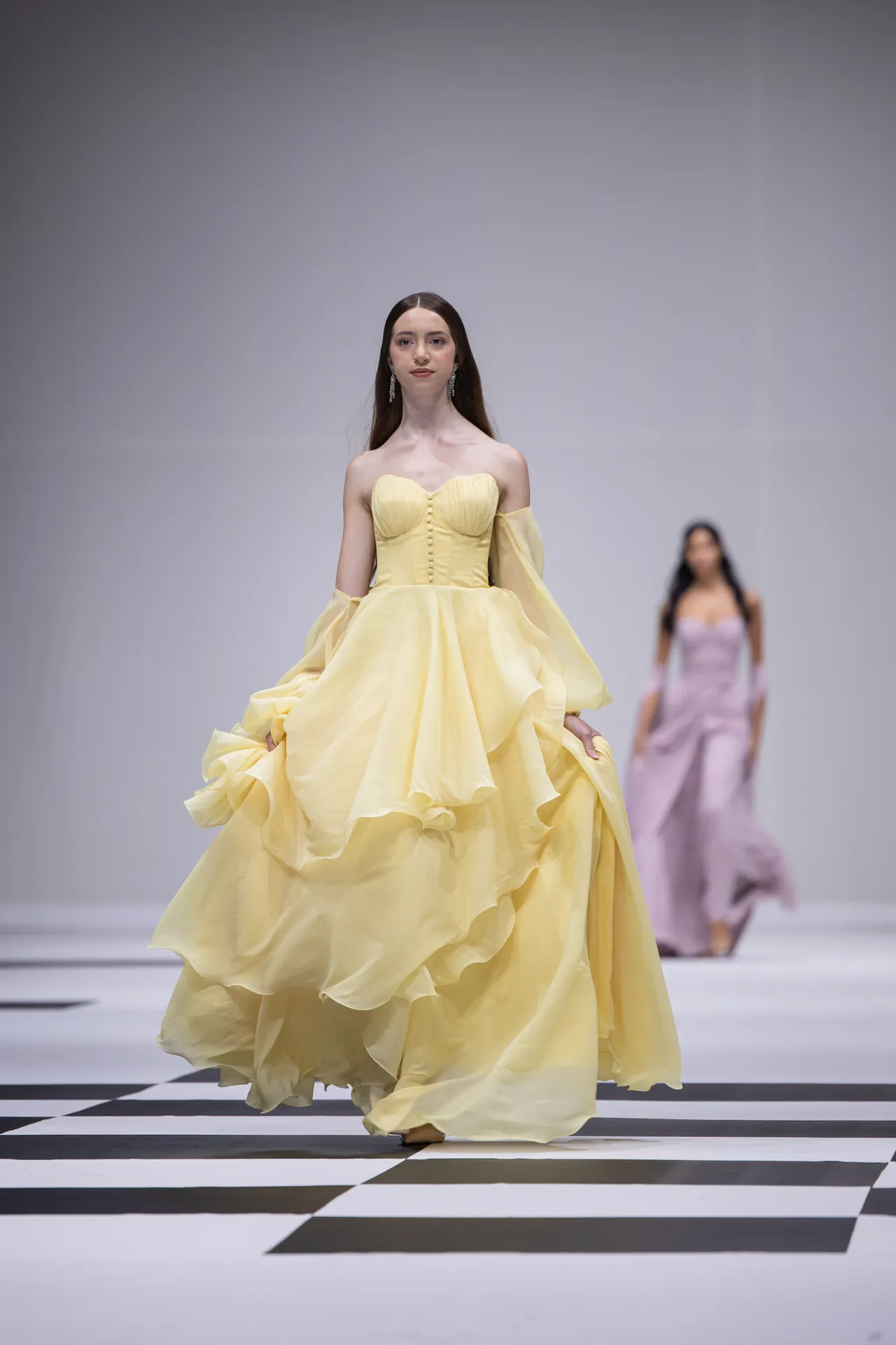 Model walks the runway in a pale yellow, off-shoulder ball gown with a corseted bodice and voluminous skirt; black-and-white checkerboard floor beneath.