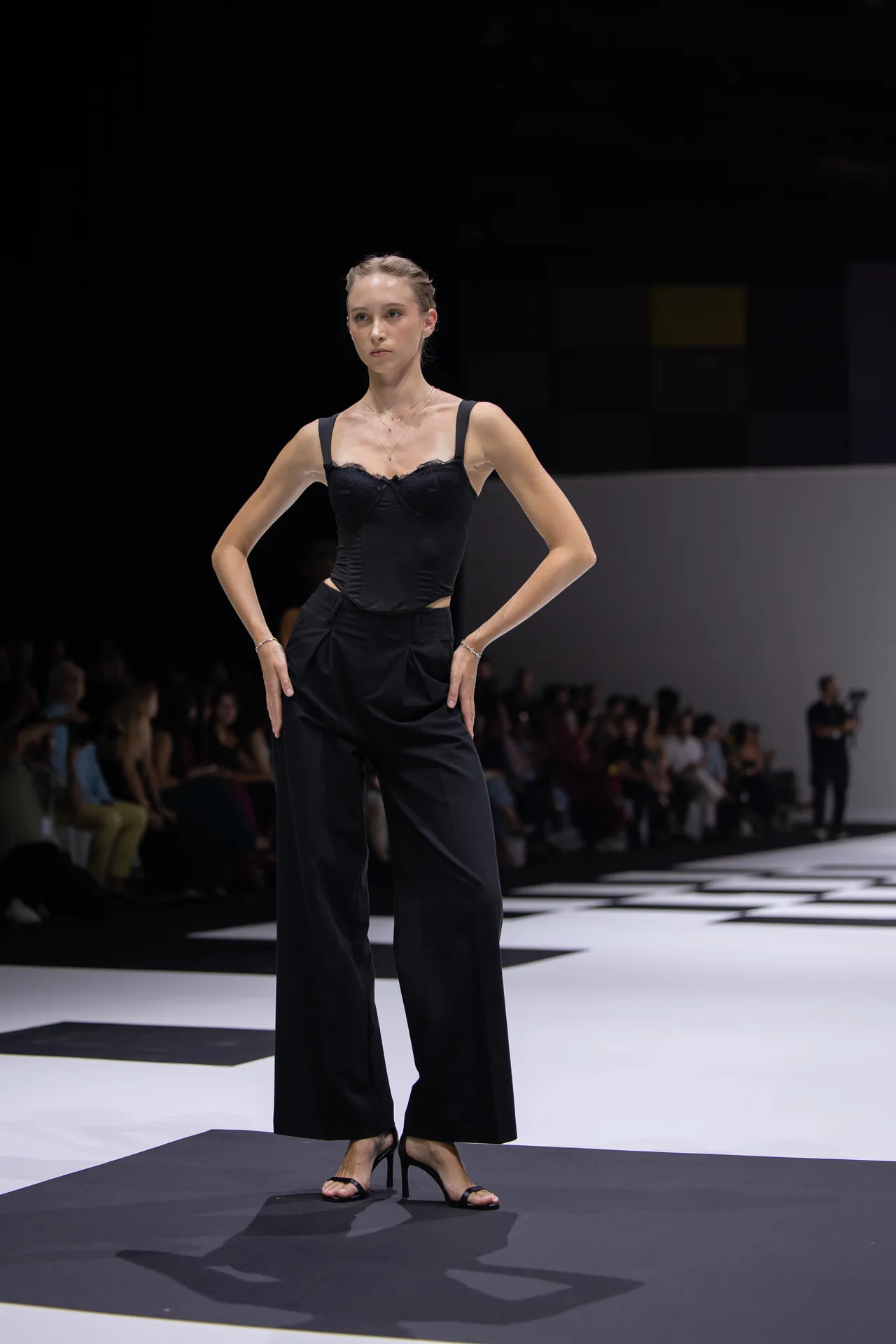 Model on a runway wearing a black, structured corset top and wide-leg pants, posing with hands on hips amid a black-and-white patterned floor and dark background.