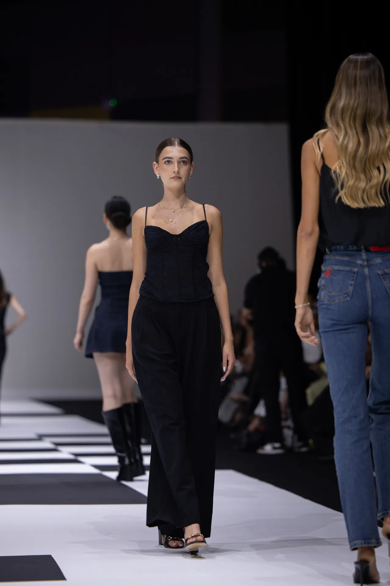 Model walks a black-clad look down a fashion runway, other models and audience in the background.